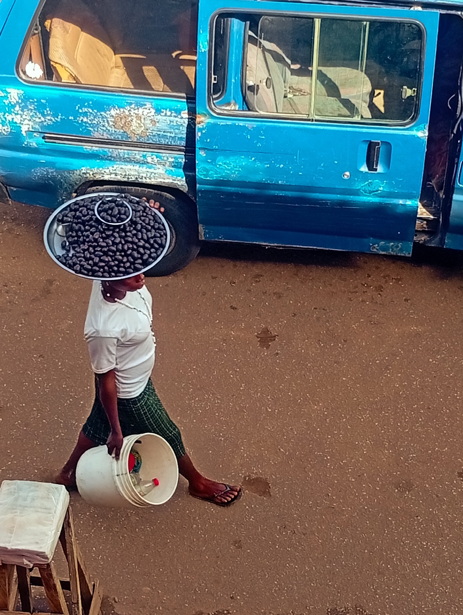 A woman walks across a dirt road in Bukuru, Nigeria, carrying a large metal basin filled with black berries on her head. She wears a white t-shirt, a green checkered skirt, and flip-flops. In her left hand, she carries a large white bucket. To her left, a wooden stand covered in white plastic wrap is visible. Behind her, a faded blue van with its doors open is parked on the side of the road. The van's interior, including seats, is visible through the windows. The ground is a dusty brown color, with scattered small pebbles and some water stains.