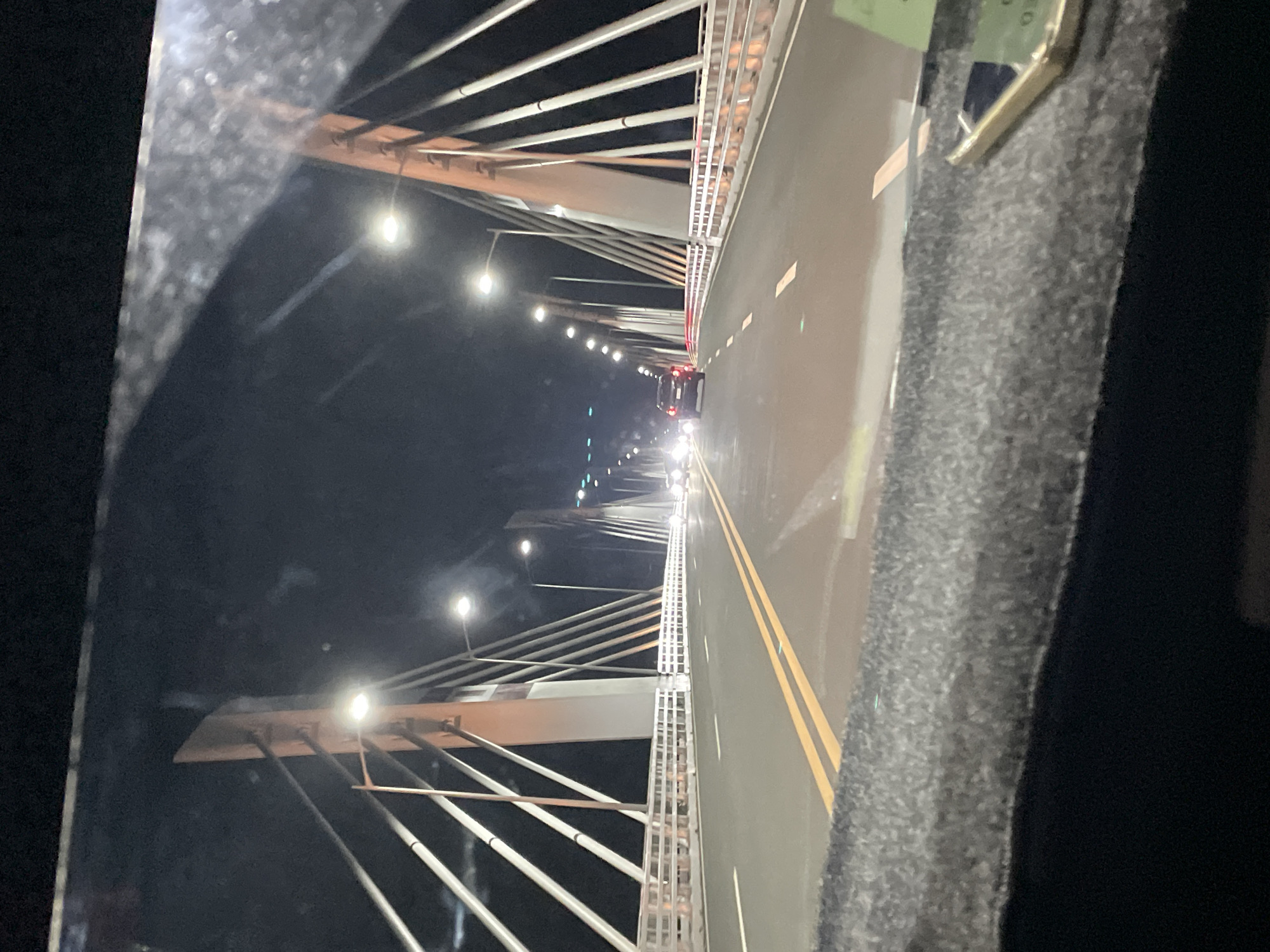 The image depicts a view from inside a vehicle traveling on a bridge at night. The bridge has a cable-stayed design with support structures visible on both sides of the road. Streetlights illuminate the bridge and road. Traffic is present, with vehicle lights visible in the distance. The camera appears to be located inside of a vehicle with interior details visible on the right and left of the frame. Given the cable-stayed bridge design, the location is highly probable to be the Davao City–Samal Bridge, in Davao City, Philippines.