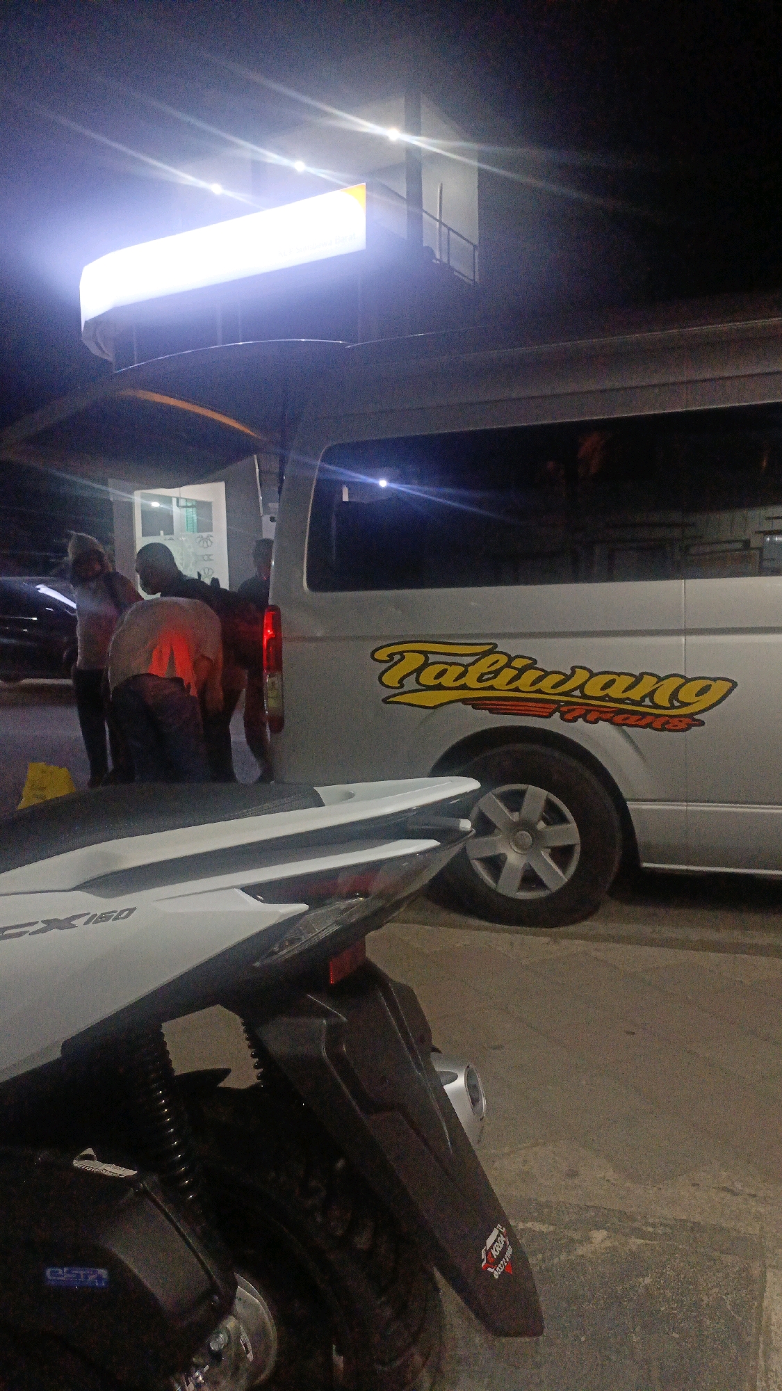 The image captures a nighttime scene in Taliwang, Indonesia. A silver van with the word "Taliwang" prominently displayed in yellow lettering on its side is parked on a paved surface. The van has a red taillight illuminated. In the foreground, the rear section of a white and black motorcycle is visible, with "GX 150" printed on its white bodywork. A sticker with red and white lettering is affixed to the motorcycle's fender. Several individuals are standing near the van, with one person in a light-colored shirt appearing to be bent over. A bright, rectangular illuminated sign hangs above and to the left of the van. The environment appears to be an outdoor area, possibly a commercial establishment or rest stop. The lighting is dim, with the bright sign and vehicle lights providing the primary illumination.