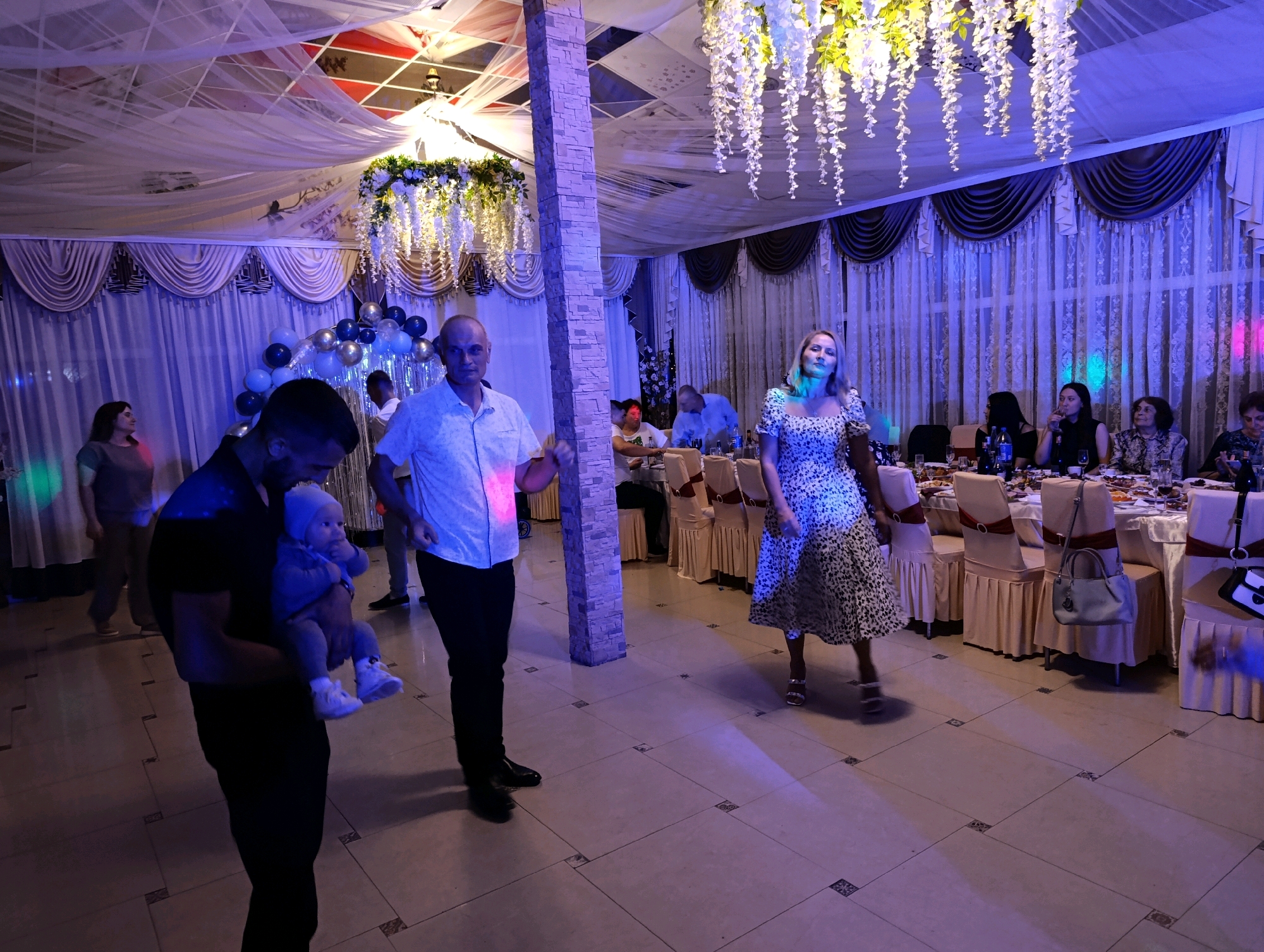 The image depicts an indoor gathering or celebration, possibly a wedding reception or a party, taking place in a venue in Korzhevo, Moldova. The room is decorated with draped white fabric from the ceiling, creating a festive atmosphere. There are also hanging floral arrangements, and strings of white lights that contribute to the ambiance. The walls are adorned with elaborate, pleated curtains in muted tones, accented with darker borders.
In the foreground, a man in a black t-shirt and dark pants is holding a baby, who is wearing a light-colored outfit and a hat. The man is facing away from the camera, his back to the viewer. To his right, a man in a white short-sleeved shirt and black pants is standing and looking towards the camera. He appears to be dancing or moving.
Further into the room, a woman in a patterned dress is walking, also appearing to be dancing or moving. She has blonde hair and is wearing heels. In the background, several tables are set up with chairs, suggesting a dining area. People are seated at some of these tables, engaged in conversation or eating. The tables are covered with tablecloths and set with plates, glasses, and bottles.
The lighting in the venue is dim, with prominent blue and pink hues cast by colored lights, creating a vibrant and somewhat moody environment. The floor is tiled with a light-colored pattern. The overall impression is of a lively event with a mix of people enjoying themselves in a decorated indoor space.