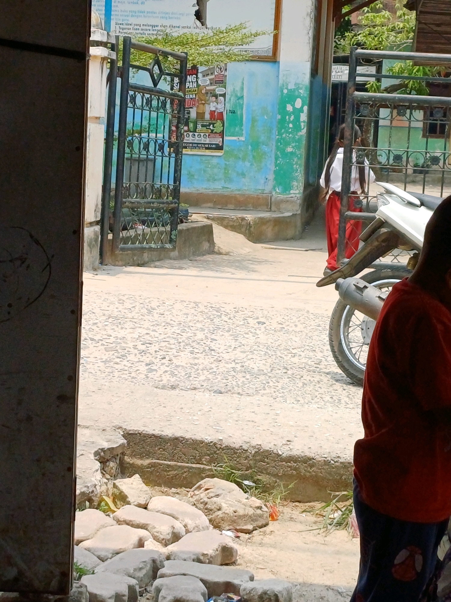 In the city of Dumai, Indonesia, a street scene unfolds outside a building with a faded teal and light blue facade. A black metal gate, partially open, stands in the foreground, revealing a path beyond. Two young girls, one in a white shirt and red pants and the other only her red pants visible, are partially obscured by the gate. The girl in the white shirt and red pants has long dark hair and is facing away from the camera, walking towards the right. A white motorcycle is parked on the paved ground to the right of the gate. In the immediate foreground, a person wearing a red shirt and dark patterned pants is visible from the waist down, facing away from the camera and towards the right. The ground is a mix of gravel and concrete, with some scattered rocks and dry grass. A poster with Indonesian text and images is affixed to the building wall behind the gate. The overall impression is a sunny day in a somewhat worn urban environment.