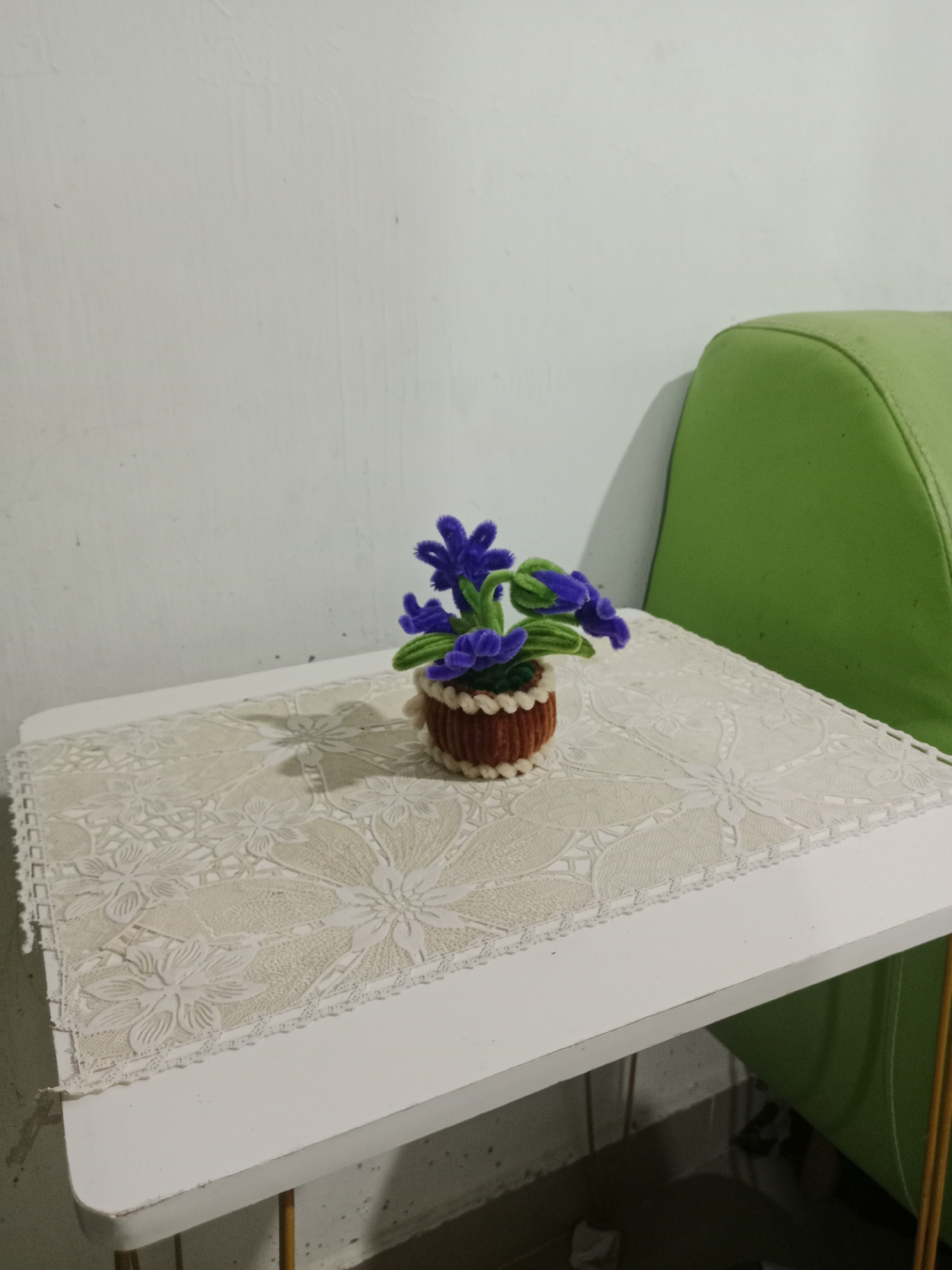 A miniature potted plant with purple flowers and green stems sits on a white table covered with a beige lace tablecloth. The table is situated next to a bright green cushioned chair. The background consists of a white wall. The scene is indoors, possibly in a home or a small shop. The image was taken in Tambakcekur, Indonesia.