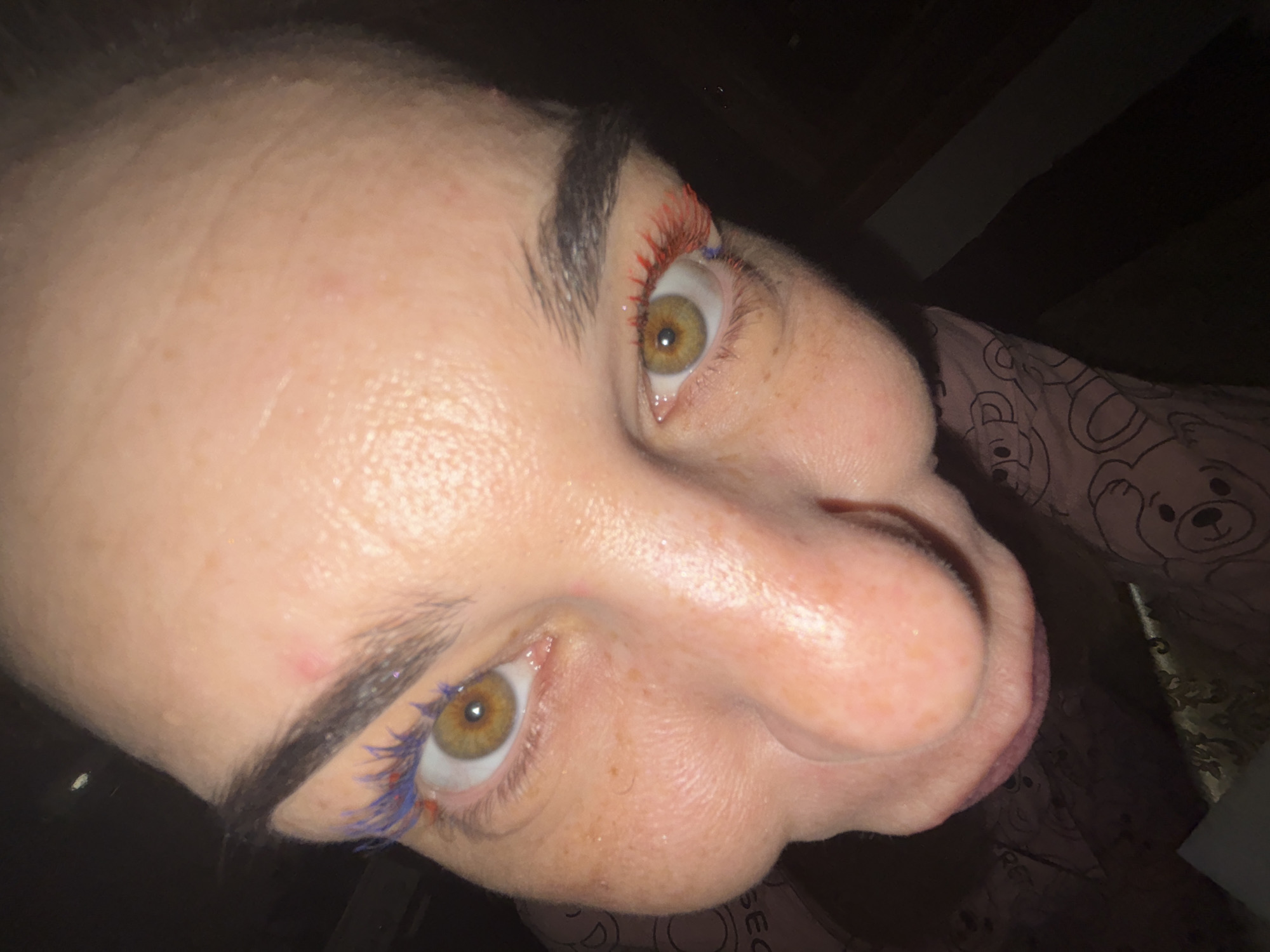 The image is a close-up, fisheye perspective of a person's face, specifically focusing on their eyes, nose, and forehead. The person has brown eyes and is wearing colorful mascara, with blue on the left eyelashes and red on the right. The skin appears to have freckles and some minor blemishes. The background is dark and out of focus, making it impossible to discern any environmental details or city location.