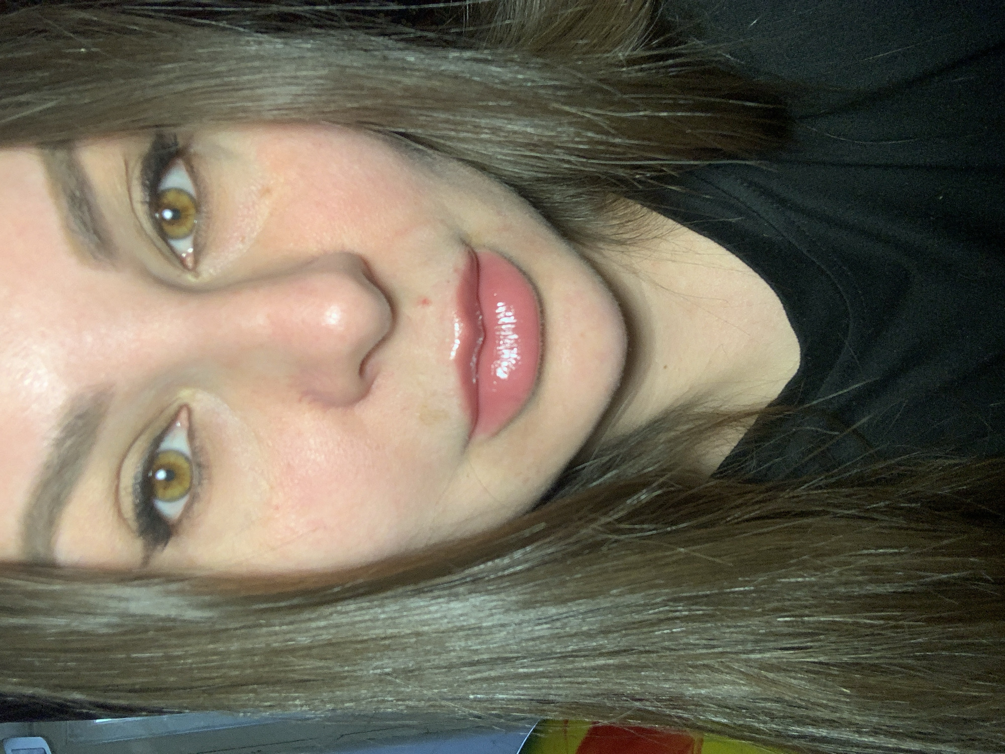 A close-up, head-on, indoor, shoulder-up selfie shows a young woman with long, dark brown hair. She is looking directly at the camera. Her eyes are a striking amber color, and she is wearing black eyeliner and mascara. Her lips are full and glossy, with a pinkish-nude lipstick. She is wearing a plain black t-shirt. The background is dark and out of focus, with hints of color on the left side, possibly indicating a colorful object or wall. The lighting appears to be artificial, casting a slight shadow on her left cheek.