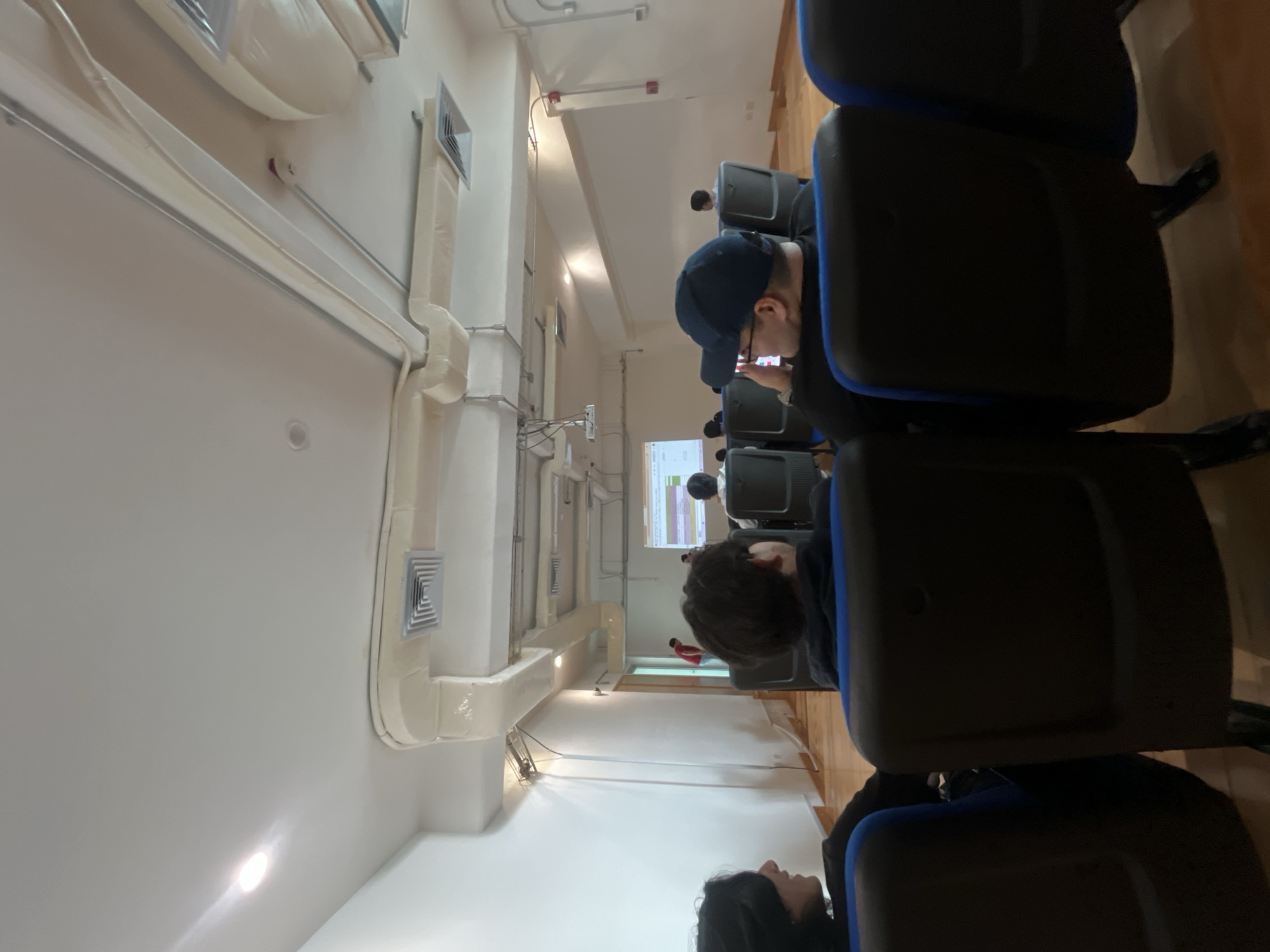 A classroom in Unknown, Unknown, has rows of black and blue chairs arranged in front of a projection screen showing a presentation. Several people are seated, some facing the screen while one wears a baseball cap and looks at a phone. The room is brightly lit, with white walls and ceiling. Ventilation ducts run along the top of the walls.