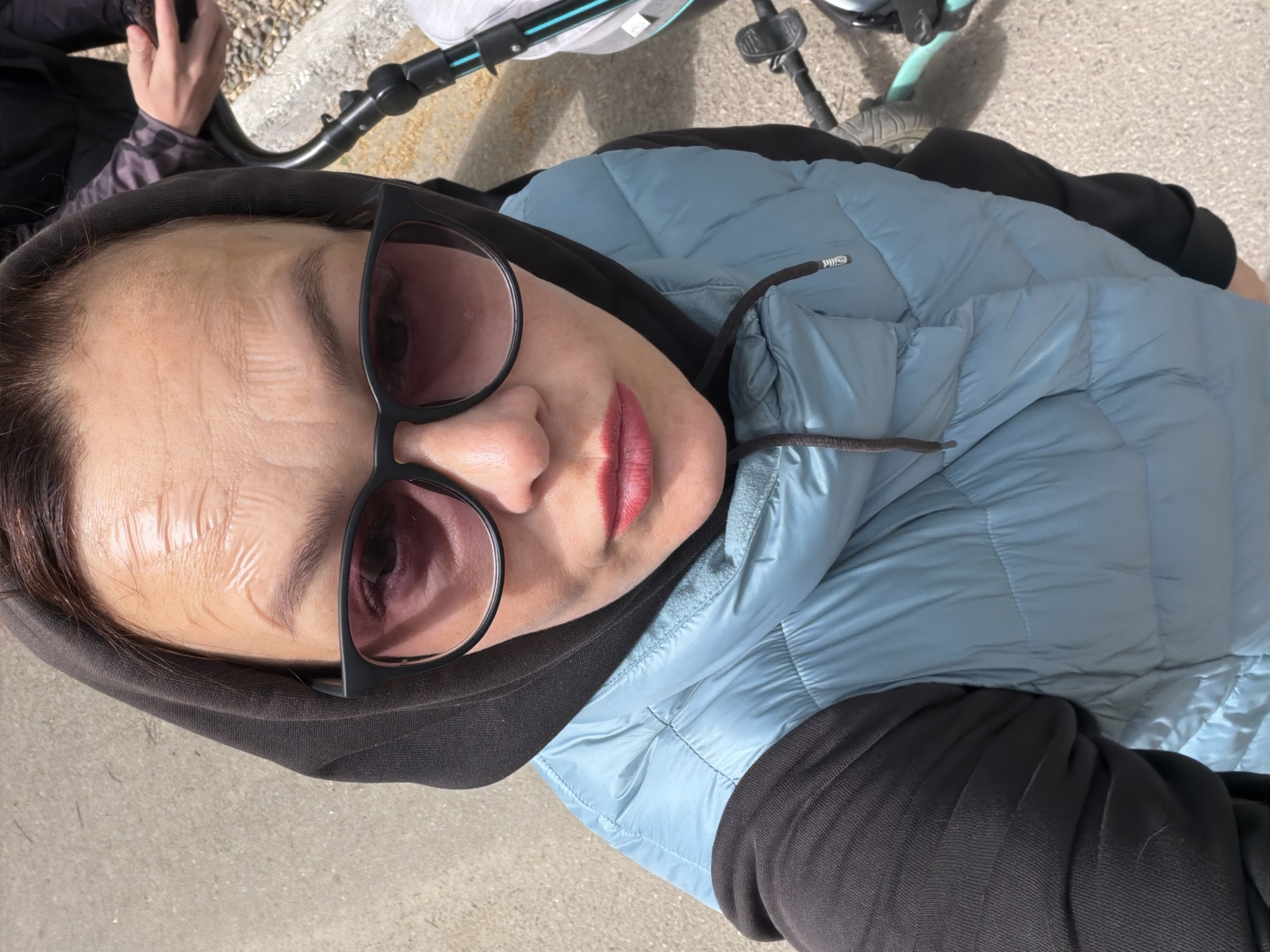 A woman wearing a black hoodie, a light blue quilted jacket, and black sunglasses is positioned at an oblique angle in the image frame. Her face is prominently featured, showing lines on her forehead, and she is wearing lipstick. Above her and slightly out of focus are components of a stroller, including parts of the frame and wheels, suggesting the woman is either pushing or standing near a stroller. The background appears to be a paved surface, likely a sidewalk or pathway. The scene is outdoors and appears to be a daytime shot. Without additional information, precise location details within the city of Chaikovsky, Russia, cannot be determined.