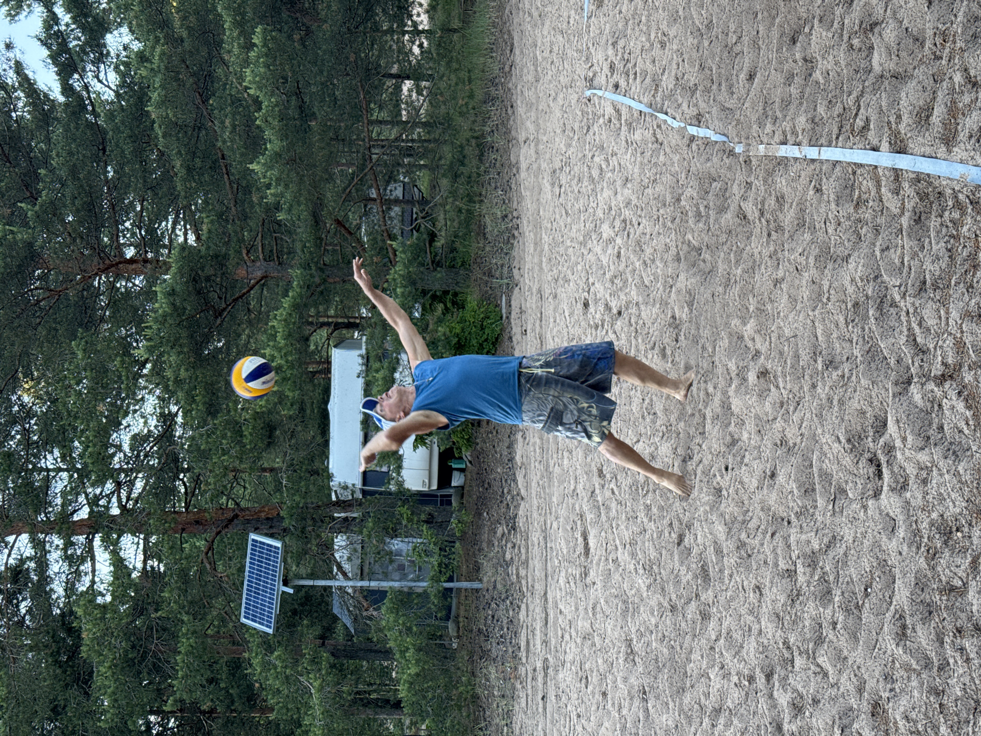 A man in a blue tank top and camouflage shorts is playing volleyball on a sandy court in Zelenaya Roshcha, Russia. He is in the process of hitting the ball, which is in the air above him. The court is bordered by a blue line. In the background, there are trees, a solar panel on a pole, and a small building. The sky is overcast.