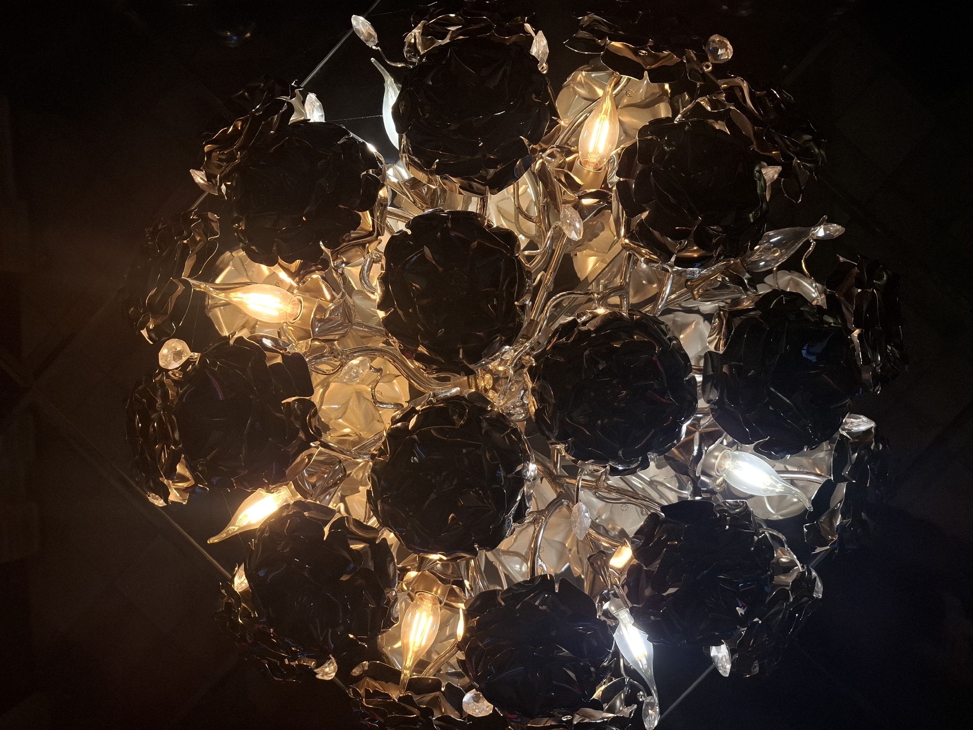 The image shows a complex chandelier hanging from a ceiling. The chandelier is spherical in shape and is composed of numerous black floral shapes. There are light bulbs interspersed among the floral shapes, emitting a warm glow. The rest of the background is dark. There is no evidence of the location being in the city of Zadonsk, Russia. There are no people visible in the image.