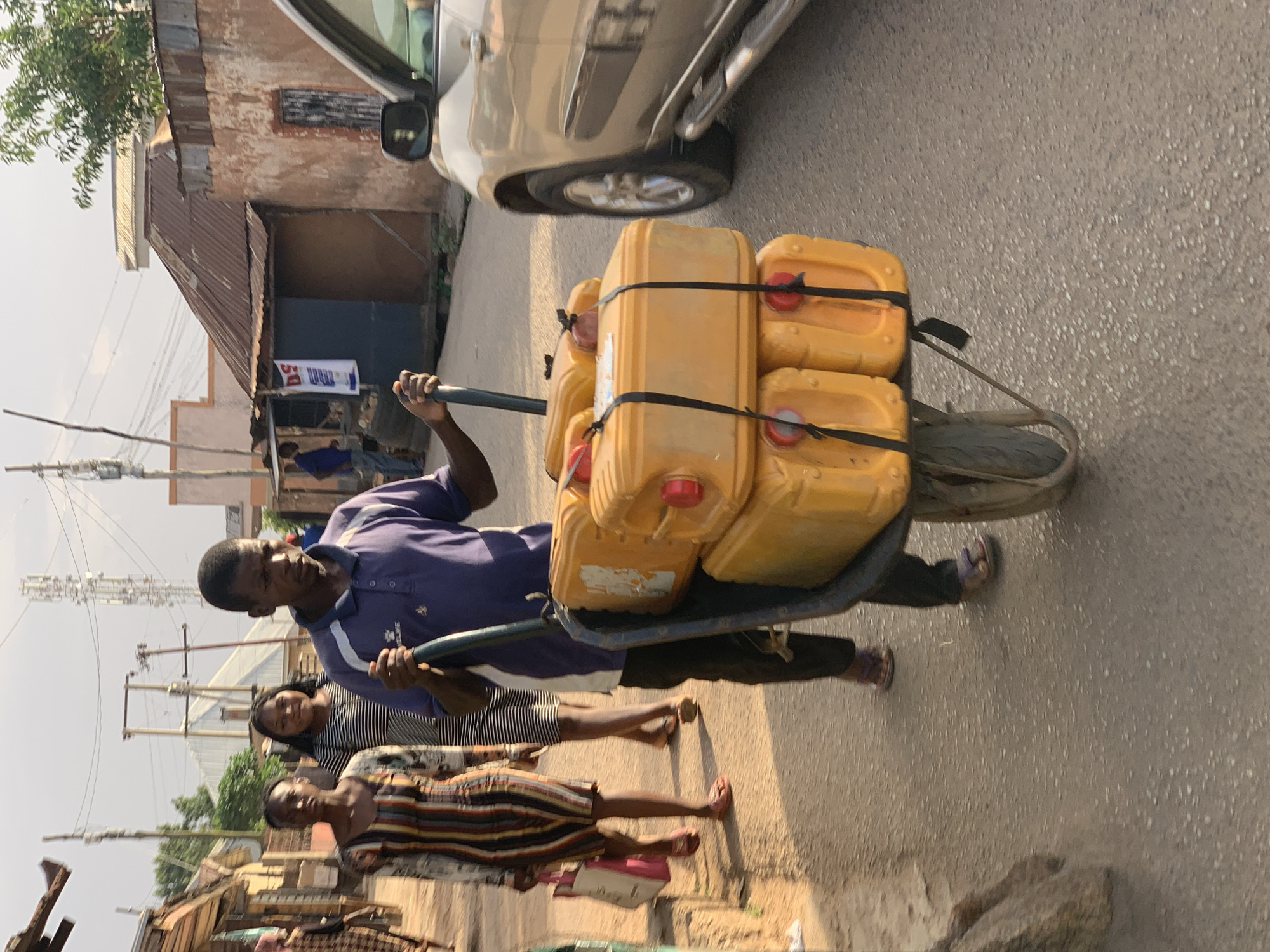 A man is pulling a wheelbarrow containing several yellow jerrycans, secured with black straps, along a street. Two women are walking alongside the man and the wheelbarrow. The scene appears to be in a residential or commercial area with buildings of varying condition, some with corrugated metal roofs. A portion of a vehicle is visible in the background. Power lines are visible overhead. The scene is illuminated by sunlight. Although I cannot confirm the exact location within Jos, Nigeria, the visual characteristics of the structures and street scene suggest a developing urban environment.