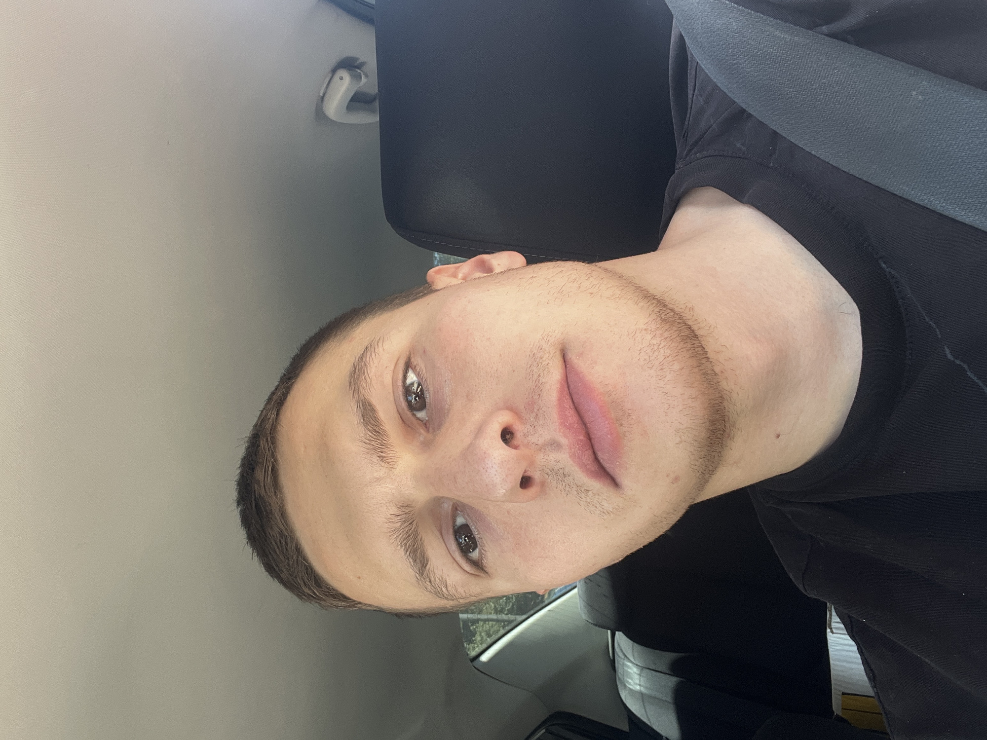 A young adult male is depicted in a close-up, tilted selfie taken from within a vehicle. He is wearing a black shirt and a seatbelt. The vehicle's interior is visible, including the headrest and ceiling. The backdrop includes an out-of-focus glimpse of greenery through the car window, possibly indicating an outdoor location. There are no definitive clues to suggest this image was taken in Burgas, Bulgaria, without further information.