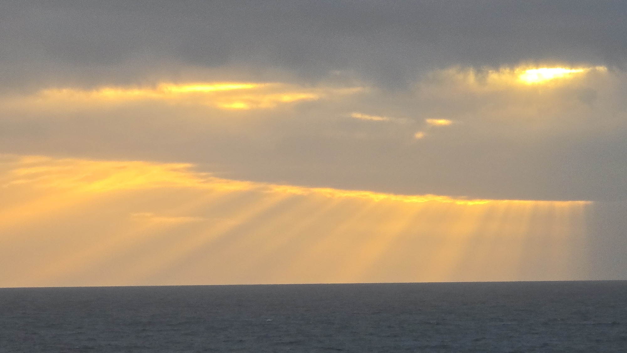 The image depicts a body of water, likely an ocean or sea, under a cloudy sky with sunlight breaking through. The sun's rays are prominently visible, extending down towards the water's surface. The location is not specified, nor is any city. No people or other identifiable subjects are visible in the scene. The environment consists solely of the sky and the body of water.