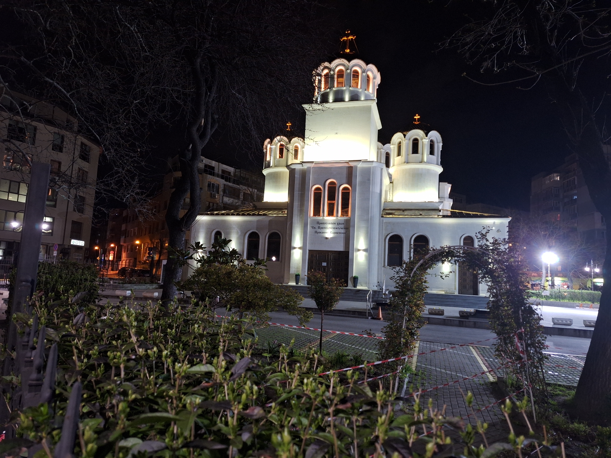 At night, the Sveti Prokopiy Varnenski Church in Varna, Bulgaria, is brightly illuminated against the dark sky. The white building, with its distinctive onion domes topped with golden crosses, is the focal point. The church is set back from the road, with a paved pathway leading to its entrance. A decorative archway covered in climbing plants frames the pathway. In the foreground, bushes and a low wrought-iron fence are visible. Red and white barrier tape stretches across the pathway, suggesting the area is restricted. Streetlights and buildings are visible in the background, indicating an urban setting. The photo is taken from ground level.