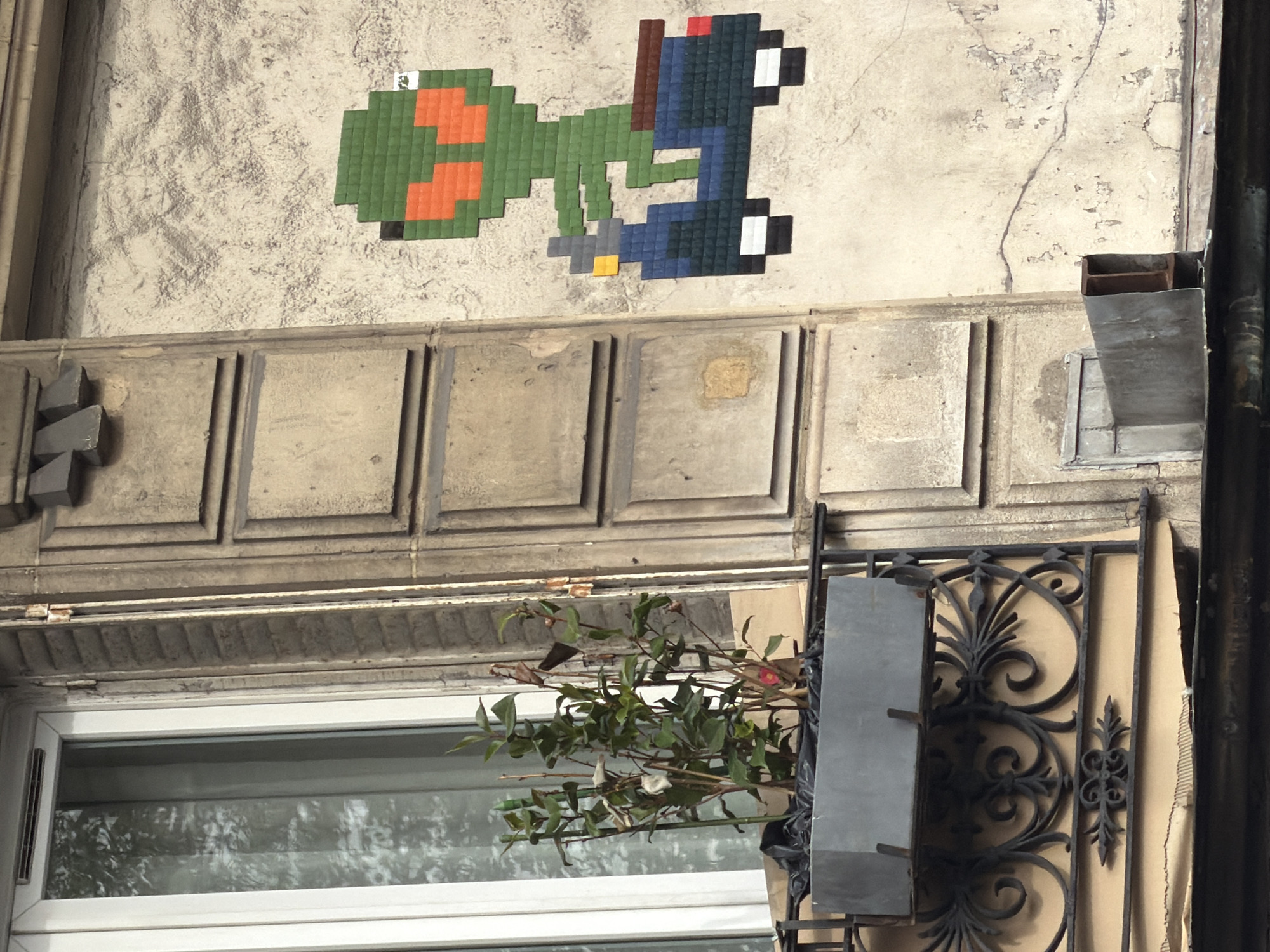 The image displays an urban outdoor scene featuring a pixelated mosaic artwork on the facade of an old building. The street art piece, stylistically associated with the artist Invader, is mounted on a textured, light-colored (possibly off-white or beige) wall. It depicts a green, pixelated creature with orange spots, resembling Yoshi, carrying a smaller blue character with a red hat, similar to Mario. A tiny white square sticker with a green pixelated icon is visible near the top left of the mosaic.
Below the artwork, the building showcases traditional architectural details, including a horizontal band of sculpted stone panels and further intricate moldings. The aged wall exhibits some cracks and wear. Beneath these architectural features is a modern white-framed window, reflecting the bright outdoor light and hints of trees or sky. In front of the window, a planter holds a lush green plant with a few reddish accents, supported by an ornate black wrought-iron structure. To the far right, a dark vertical pipe runs down the building, and several metallic fixtures, possibly utility vents or covers, are attached to the wall. The bright illumination indicates daytime conditions.