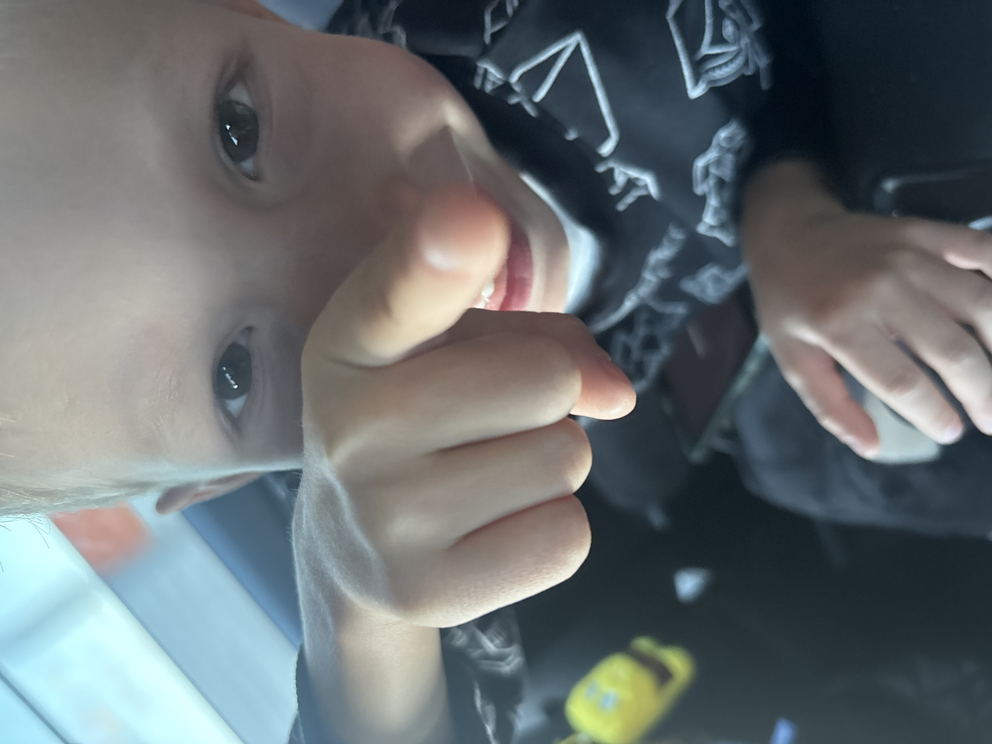 In the city of Ramezhki, Russia, a close-up shot captures a young child's face, with their right index finger prominently extended towards the camera. The child's eyes are open and focused, and their mouth is slightly agape, revealing teeth. They are wearing a dark-colored hoodie with a white graphic pattern. The background is blurred, suggesting the photo was taken inside a vehicle, possibly with sunlight streaming in from a window, casting a bright, blueish hue. A yellow object is partially visible in the lower left corner, and a black surface, likely part of the car's interior, dominates the lower half of the frame. The child's hand is in sharp focus, positioned in the foreground, emphasizing the gesture of pointing.