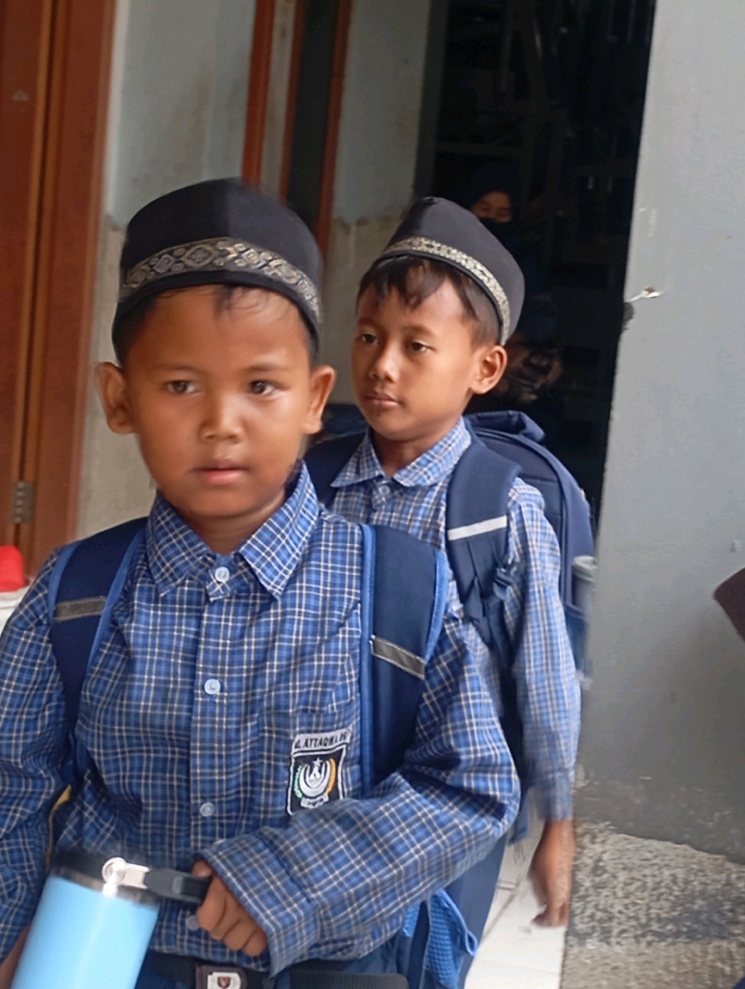 Two young boys, likely students, are the focus of this image. The boy in the foreground is wearing a black peci (a brimless cap) and a blue and white plaid collared shirt. He has a blue backpack on and is holding a light blue water bottle. The boy behind him is also wearing a peci and a similar blue and white plaid shirt, with a blue backpack. They appear to be in a doorway or hallway, with a textured wall on the right and a wooden door on the left. The environment suggests an indoor or semi-outdoor school setting, potentially in Babelan, Indonesia, given the attire and general appearance.