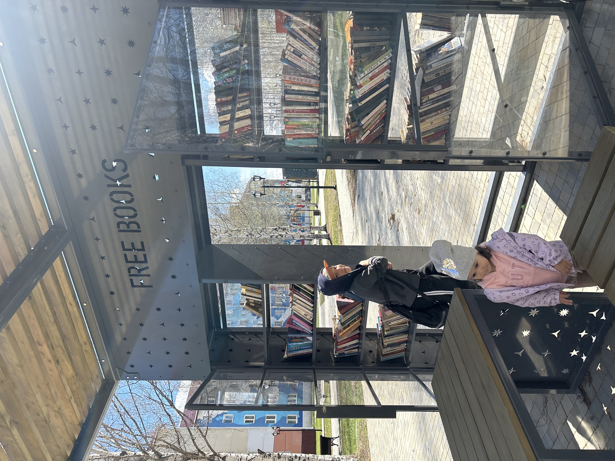 Two children, one dressed in a gray hat and black jacket, and the other in a purple jacket and gray hat with a silver heart, are positioned inside a small, public library or book exchange in what appears to be a park or outdoor area in Uray, Russia. The library features shelves stacked with books and is enclosed with glass panels and gray framework, adorned with star-shaped cutouts. The sign "FREE BOOKS" is visible. The children appear to be sitting or resting on a bench inside the structure. Outside the library, there are paved walkways, grass, trees without leaves, street lights, and colorful buildings, indicative of a residential or mixed-use area in the city.