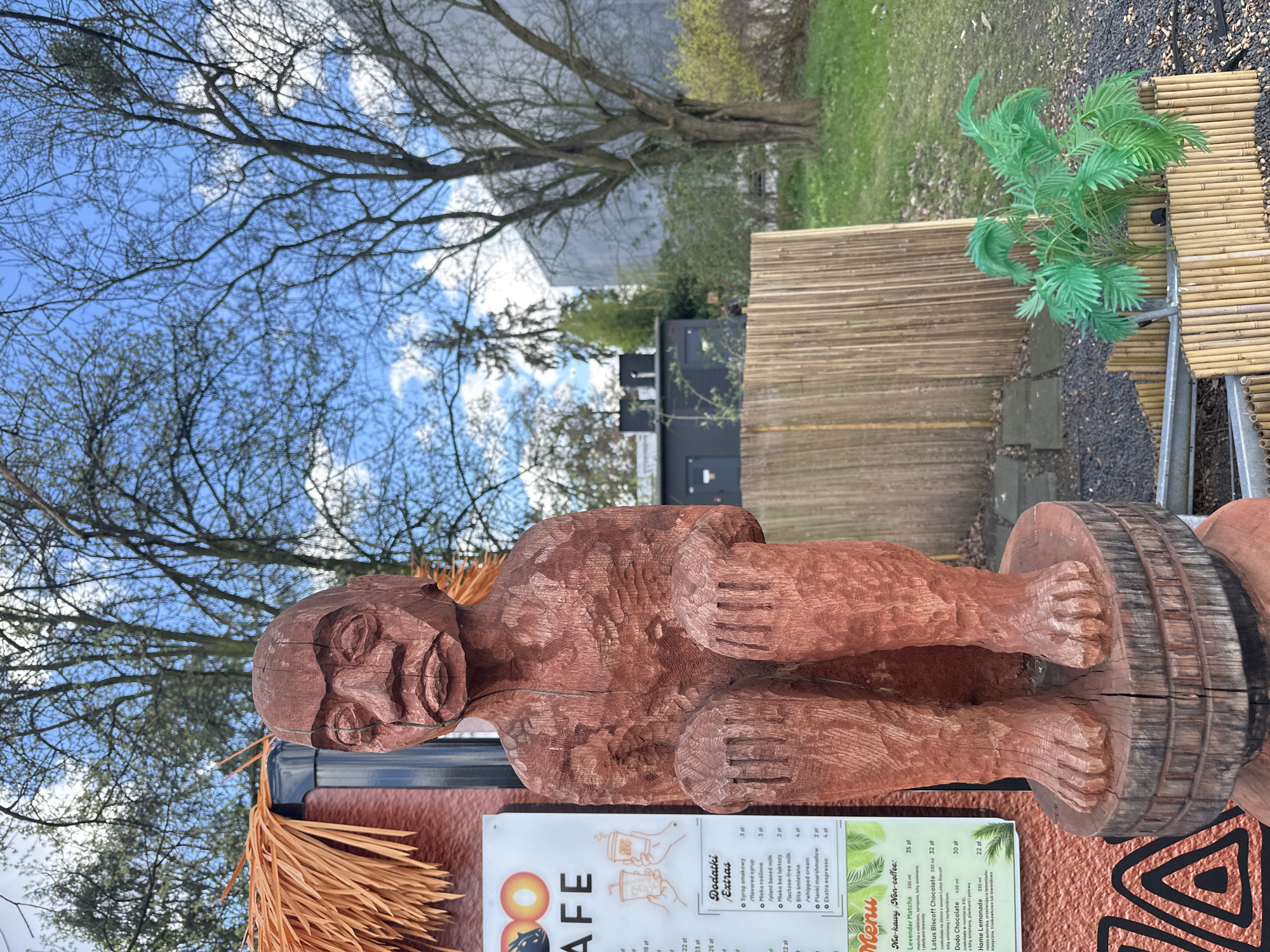 The image shows a wooden statue of a squatting figure on what appears to be a barrel. It stands next to a menu board for a cafe, with text in Polish. A faux tiki theme is suggested by the straw fringe. The background includes trees, a building, and a blue sky with clouds. The location appears to be an outdoor cafe setting, possibly in Wrocław, Poland, based on the menu’s currency in Polish złoty. A faux plant sits on a bamboo table.