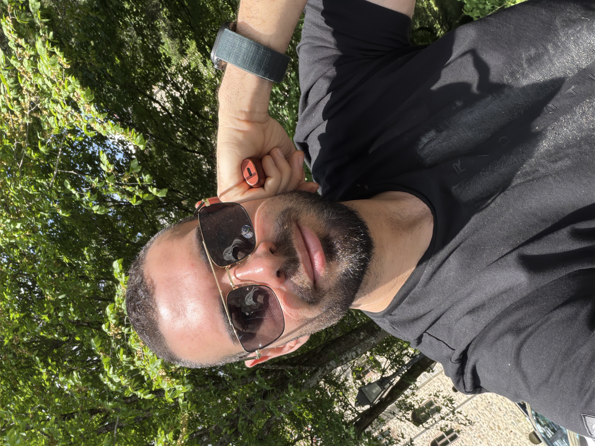 A man with a dark beard and sunglasses is shown in a close-up, selfie-style photograph. He wears a black t-shirt and a dark green smartwatch. He holds a small, orange, cylindrical object in his hand. The background shows green foliage, possibly trees. Buildings are visible in the background, consistent with the architecture of the Rilski Manastir monastery in Bulgaria. The photograph is taken outdoors in daylight.