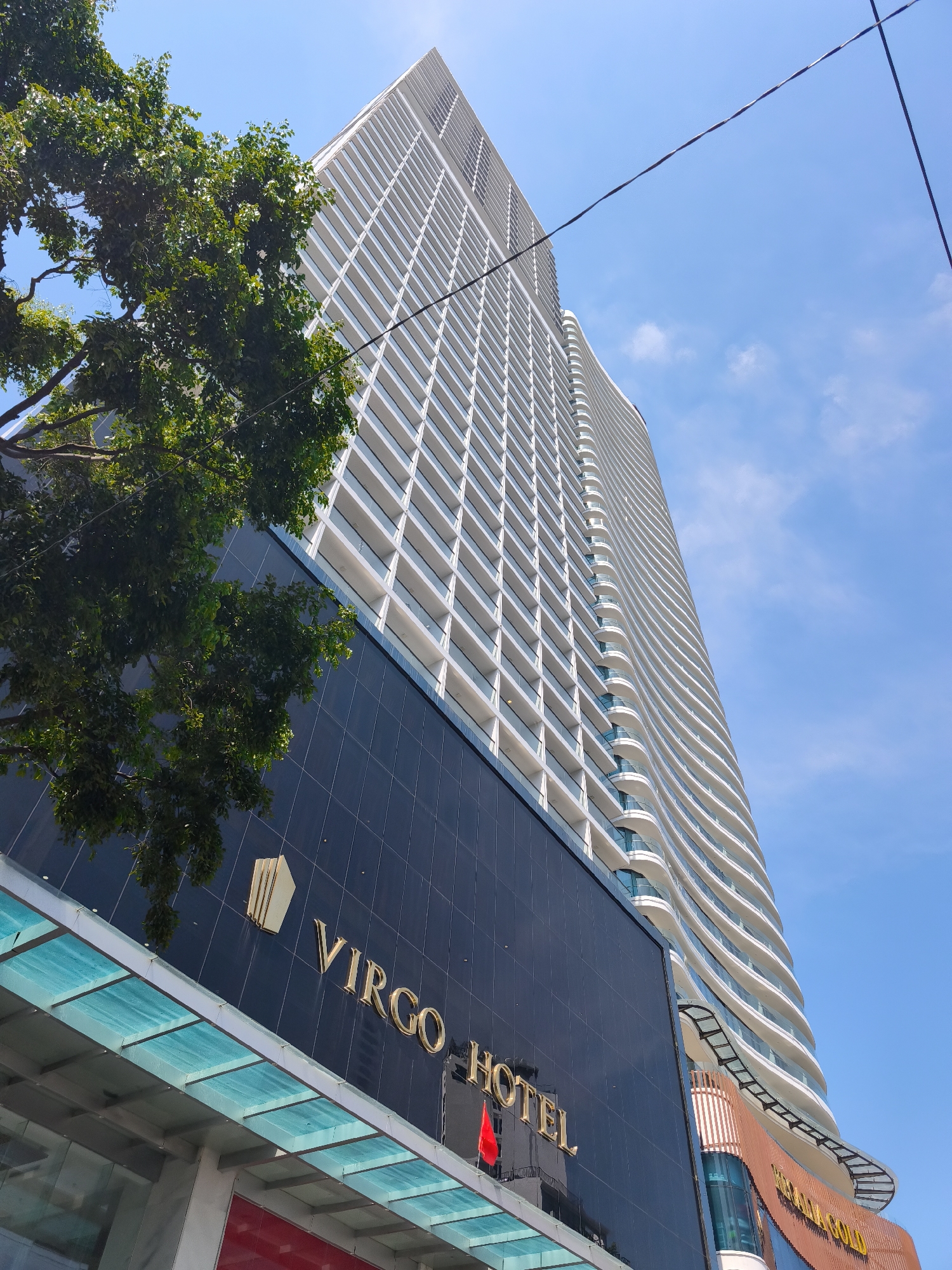 A low-angle, full-shot photograph captures the exterior of the Virgo Hotel in Nha Trang, Vietnam, under a clear blue sky with scattered clouds. The building, a modern high-rise, is predominantly white with a series of curved balconies that give it a unique architectural style. The lower portion of the building features a dark, reflective facade with the golden inscription "VIRGO HOTEL" prominently displayed, along with a golden emblem. A lush green tree with dense foliage partially obstructs the view of the building on the left side of the frame. Overhead, dark electrical wires are visible against the sky. The building's lower levels are lined with glass awnings, and the warm tones of the lower facade contrast with the cooler tones of the upper floors. The scene depicts a daytime view of the hotel.