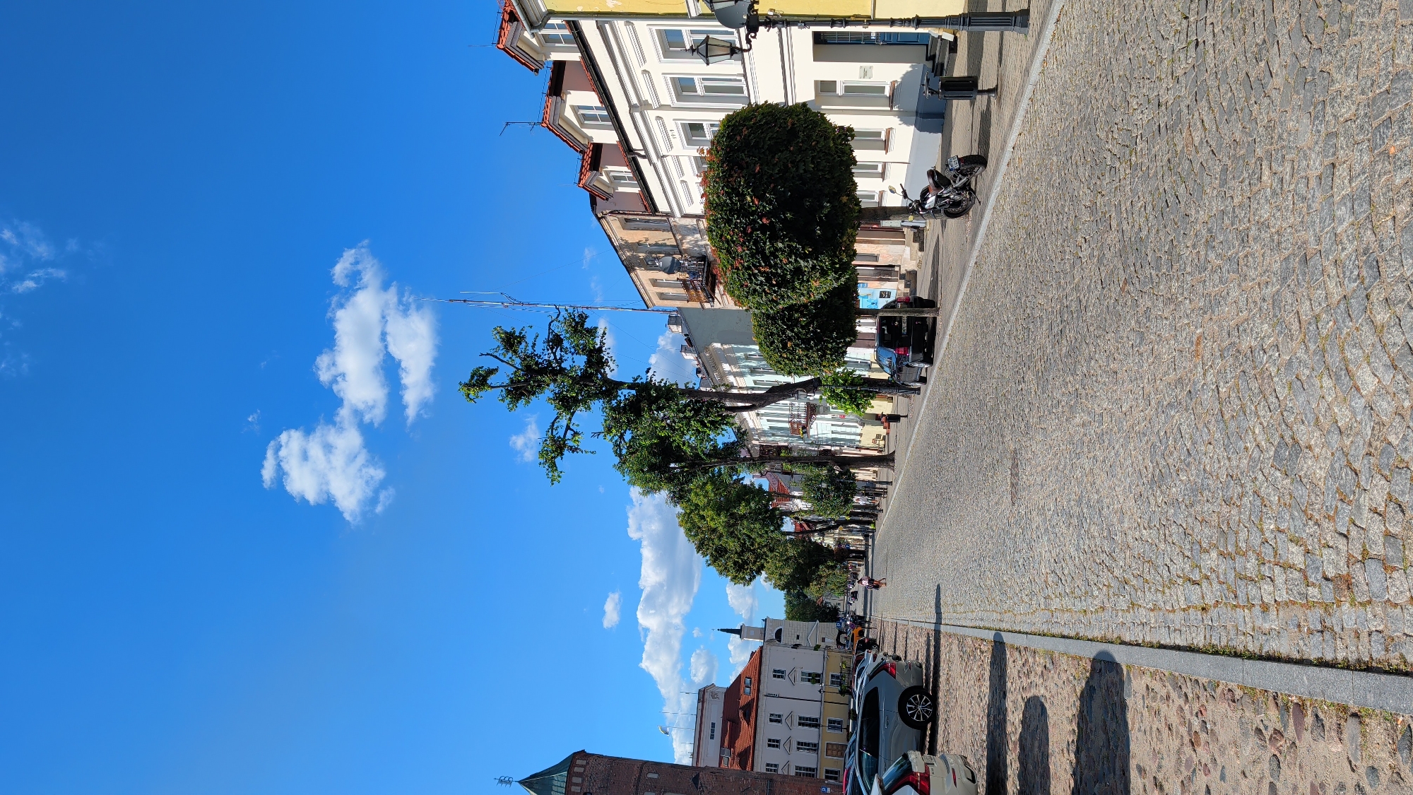 This is a vertical, eye-level, full shot of a cobblestone street in the city of Pułtusk, Poland, on a bright, sunny day with a clear blue sky and scattered clouds. The street is lined with buildings of varying architectural styles, painted in muted colors, and features mature trees with green foliage.
On the left side of the street, parked cars are visible along a stone curb. Further down the street, a historic brick tower with a green roof is prominent. A church spire can be seen in the distance.
The street itself is paved with cobblestones, and a single person is walking down the middle of the street. On the right side of the street, more buildings are visible, some with balconies. A motorcycle is parked on the sidewalk. Streetlights and trash receptacles are also present. The overall scene depicts a peaceful urban environment.
