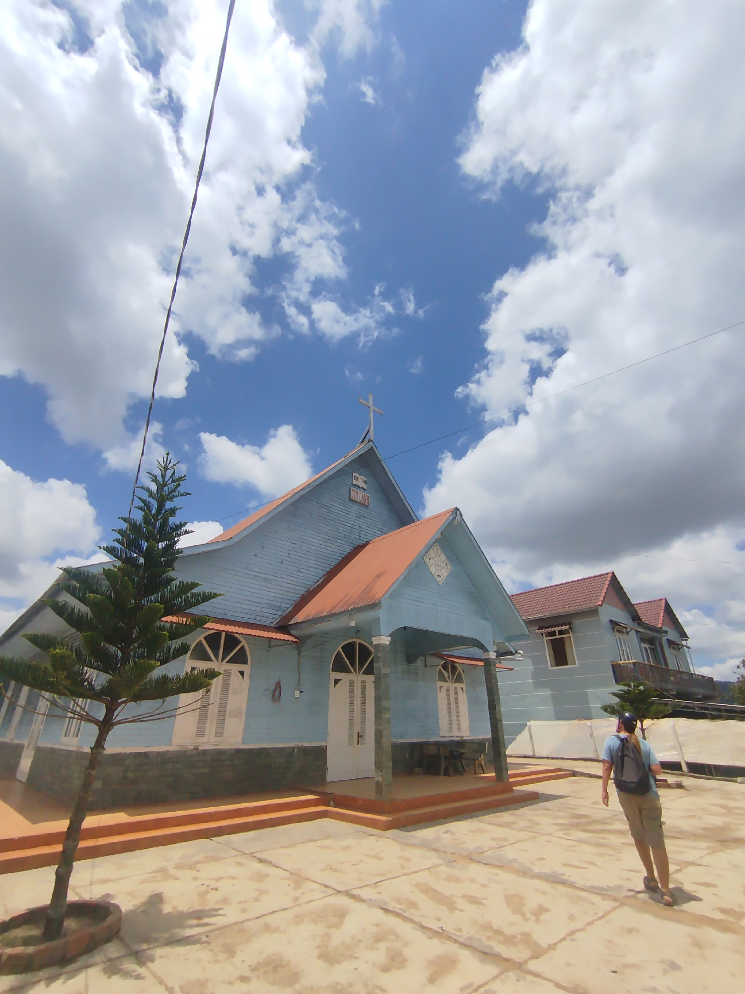 In Thôn 6, Vietnam, a light blue church with a rusty-orange roof stands under a partly cloudy sky. A small evergreen tree with sparse foliage is planted to the left of the church. In the foreground, a person wearing a blue t-shirt, khaki shorts, a backpack, and a blue helmet is walking away from the camera on a paved courtyard. The courtyard is made of concrete tiles and shows signs of wear. The church features arched windows with white shutters, and a cross is visible on the roof. To the right of the main church building, another building with a similar roof and light blue siding is partially visible, with multiple windows. A power line stretches diagonally across the sky from the top left to the bottom right of the frame.