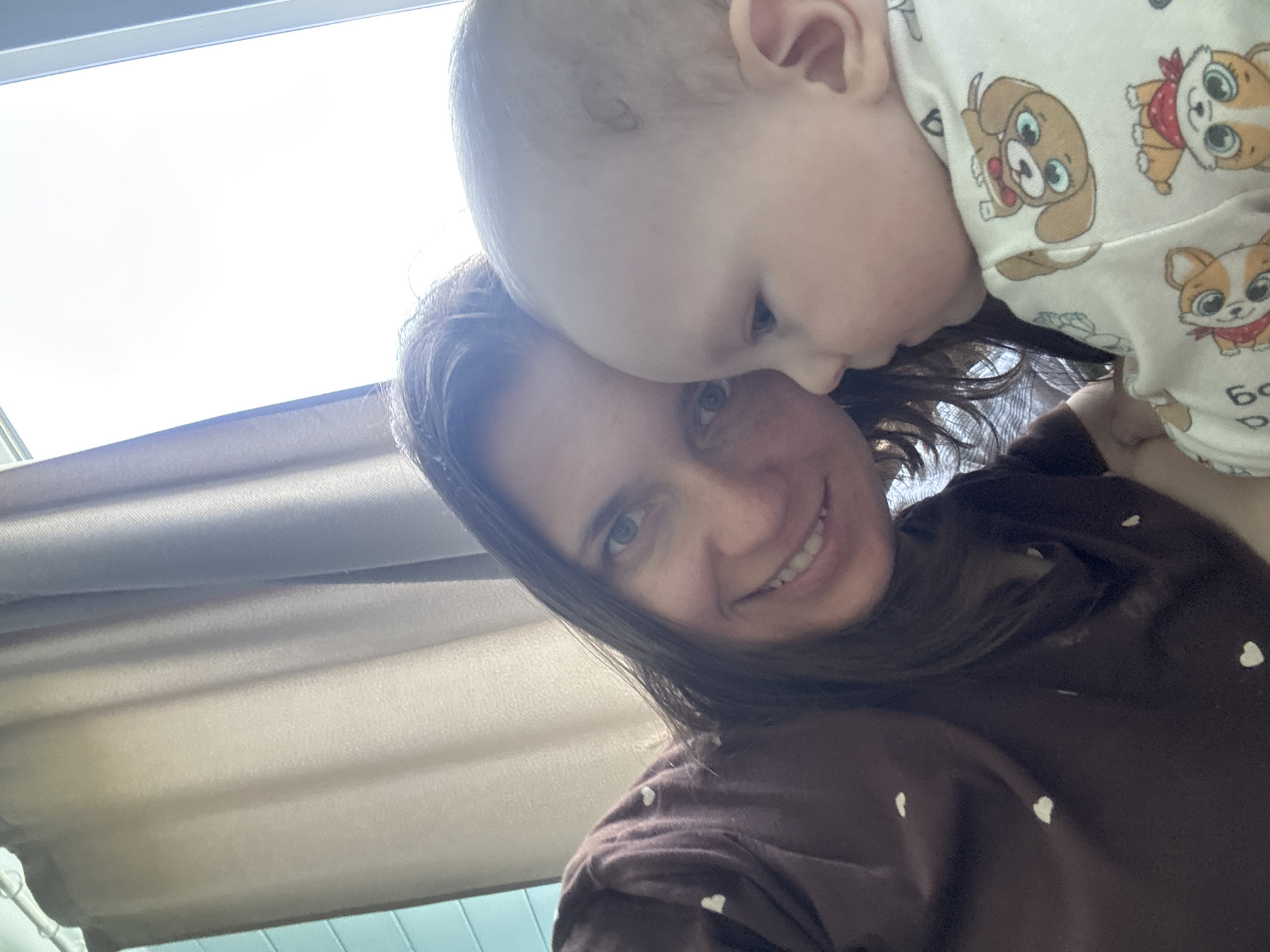 This is a close-up, angled selfie featuring an adult woman and a baby, capturing a tender moment between them. The woman, positioned in the lower right half of the frame, has long dark hair and light eyes, and is smiling warmly towards the camera. She is wearing a dark, possibly brown, top adorned with small white heart patterns. The baby, with fair skin and light hair, is seen from the side, leaning down towards the woman's head, their face partially obscured as they look towards the woman. The baby is dressed in a light-colored top patterned with various cartoon dogs: some are brown with red bandanas/collars, and others are lighter orange/tan with similar red accessories. Visible on the baby's clothing pattern, near the bottom right, are Cyrillic letters, including "Бо" and a partial "р" or "л".
The background on the top left is an extremely bright, overexposed white area, suggestive of direct sunlight or a clear sky seen through a window or sunroof. Below this, and extending to the right, is a light beige, textured surface, possibly a car's headliner or an indoor blind. A small section of light blue, possibly tiled, surface is visible at the very bottom right. The overall impression is one of affection and joy, illuminated by bright daylight. The location context for this image is Раздолье, Russia.