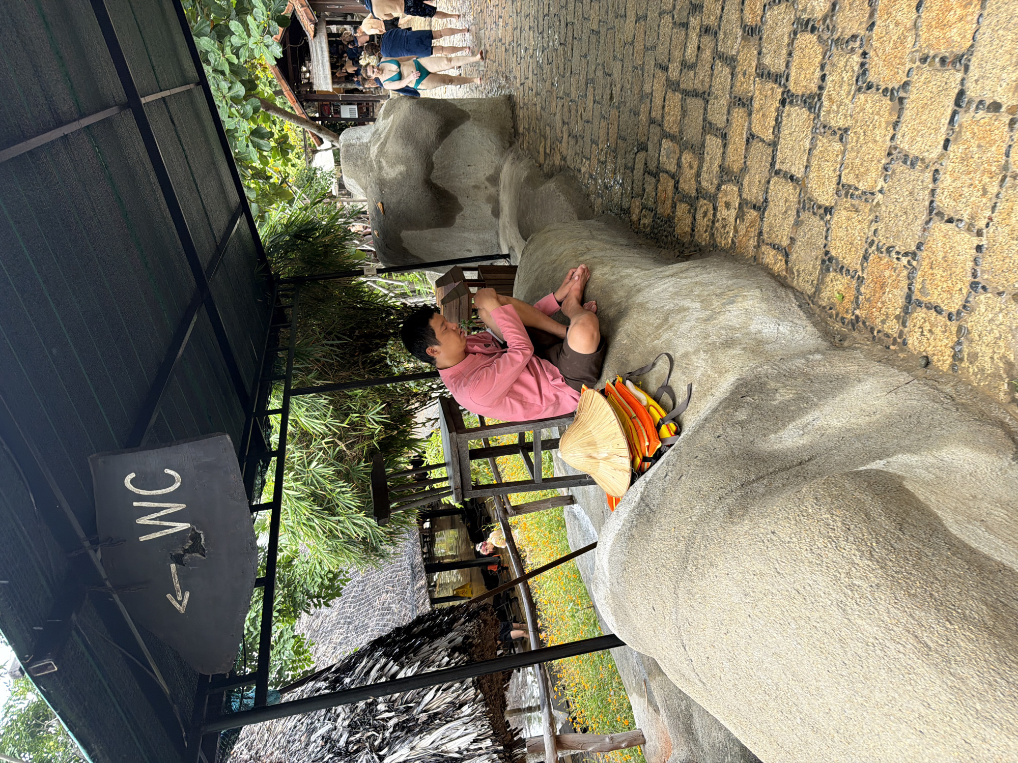 A young man in a pink shirt and brown shorts sits on a large, rough-textured rock on the left side of the frame. He is looking down at something in his hands. Beside him on the rock are a conical straw hat and several orange and yellow life vests. Behind him, there is a thatched roof structure and some greenery. A sign indicating "WC" with an arrow pointing left hangs from the overhang above. The ground to the right of the rock is a wet, cobblestone path. Further back on the right, several people in swimwear are walking. The scene appears to be outdoors, possibly at a resort or a public area with facilities, in Nha Trang, Vietnam.
