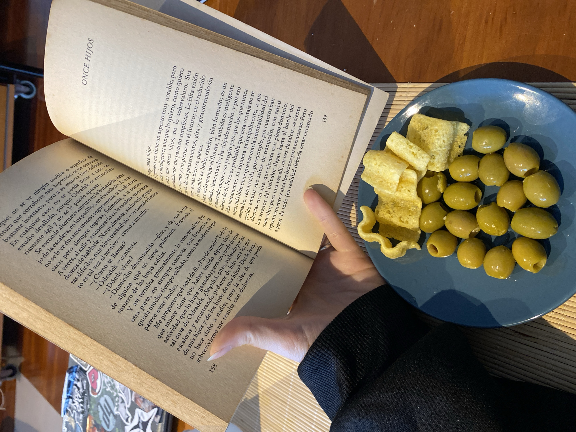 A person is holding open a book titled "ONCE HIJOS" (Eleven Sons), written in Spanish. The left page is numbered "158", and the right page is numbered "159". Next to the book, on a light blue plate, are green olives and pale, irregularly shaped, fried snacks. The person is wearing a black long-sleeved shirt or sweater. The book and the plate are on a surface with a light brown, woven texture. The location is unknown, as the image does not provide any information to determine that it is Gijón/Xixón, Spain.