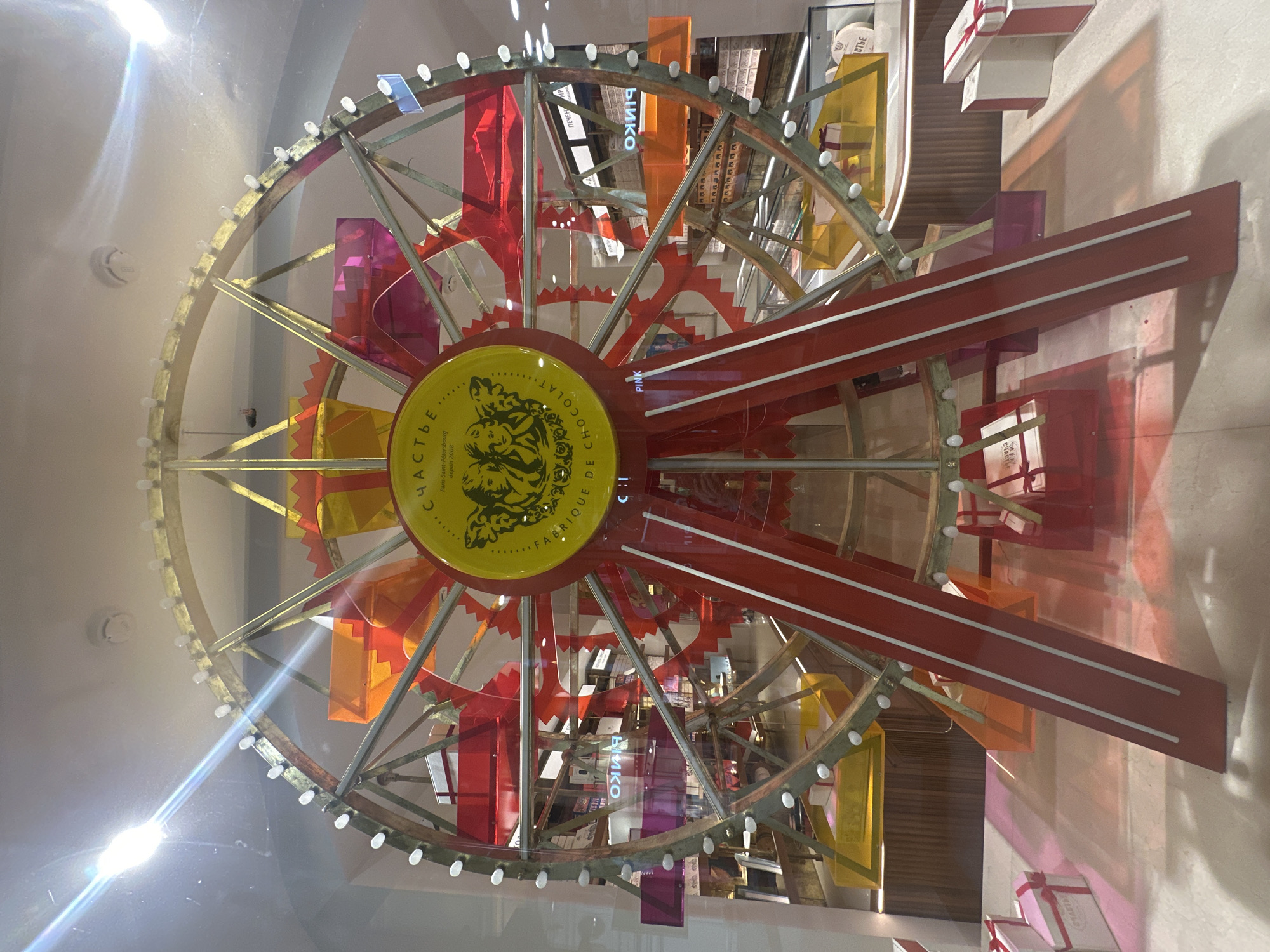 The image shows a display window, likely for a shop, featuring a large, decorative Ferris wheel. The Ferris wheel is red and gold, with clear plastic gondolas containing what appear to be gift boxes. The central medallion on the Ferris wheel has text and graphics, with the prominent word "СЧАСТЬЕ" (meaning "happiness") in Cyrillic. The background shows a retail environment with shelves and products, including some with the "PINKO" logo visible. Gift boxes are also displayed on the floor in front of the Ferris wheel. The scene is illuminated by bright lights, one of which is causing a strong glare in the upper left corner of the image. The overall impression is a festive and inviting display. While the specific location in Moscow is not identified, the presence of Cyrillic text and Russian branding suggests it is indeed in Moscow, Russia.