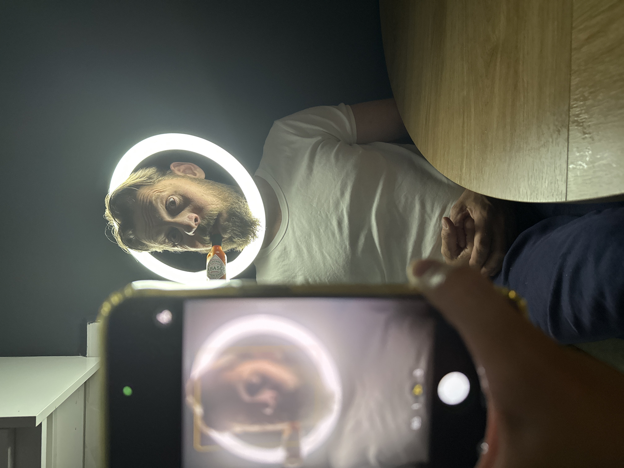 A person is taking a photo of another person with a smartphone. The person being photographed is illuminated by a ring light, and a bottle of Tabasco hot sauce is positioned in front of them. The background includes a dark wall, a light-colored shelf or table, and the edge of a wooden table. The location of the scene is not discernible from the image.