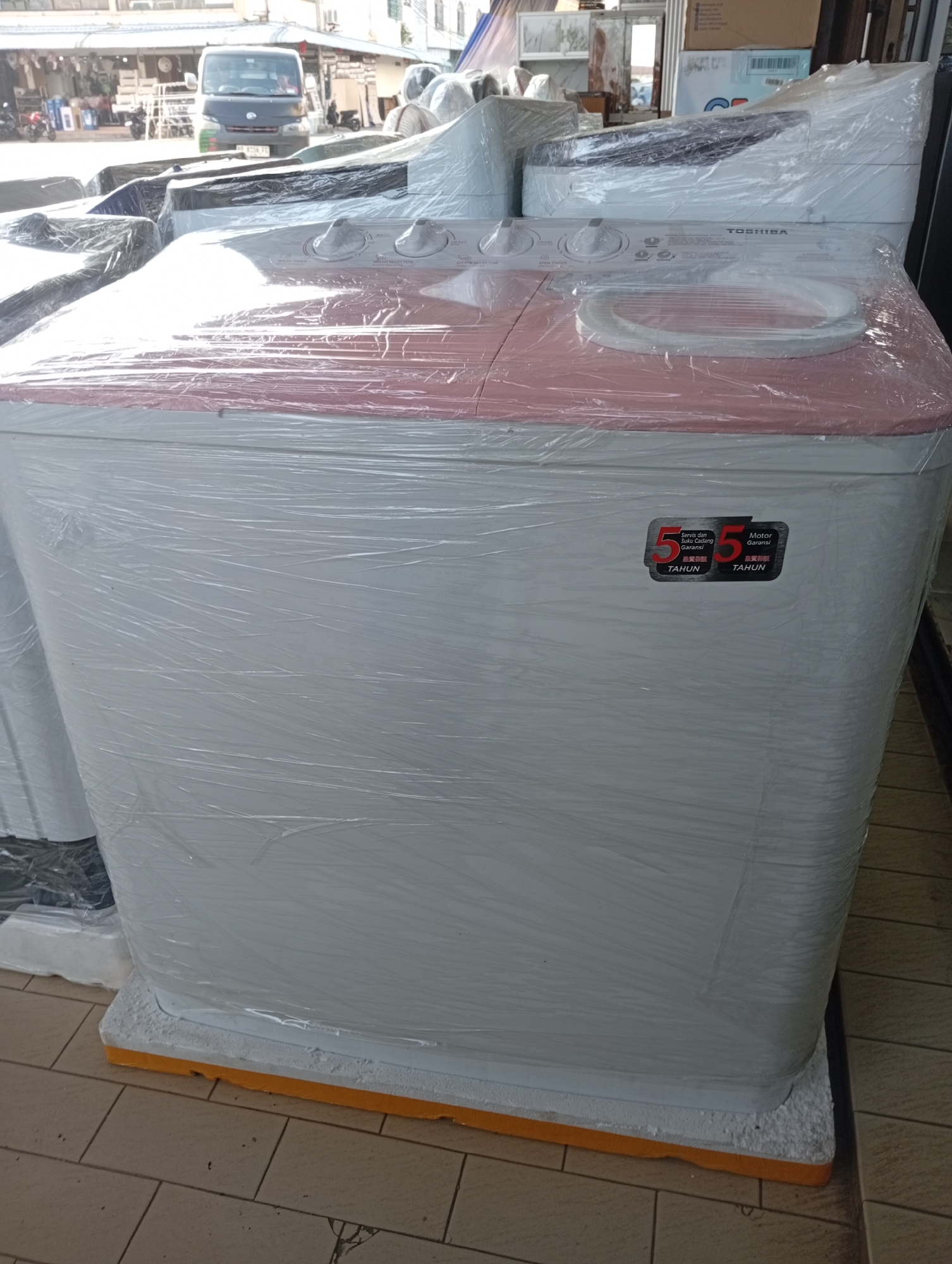 The image shows a new Toshiba twin-tub washing machine, still in its protective plastic wrapping, displayed outdoors on a tiled floor. The washing machine is white with a pink lid and features a label indicating a 5-year warranty for service and a 5-year warranty for the motor. In the background, there are other appliances, a van, and a storefront, suggesting the location is a shop or marketplace. Given the context and the presence of a van with a license plate that appears to be Indonesian, the scene is likely in Batam, Indonesia.