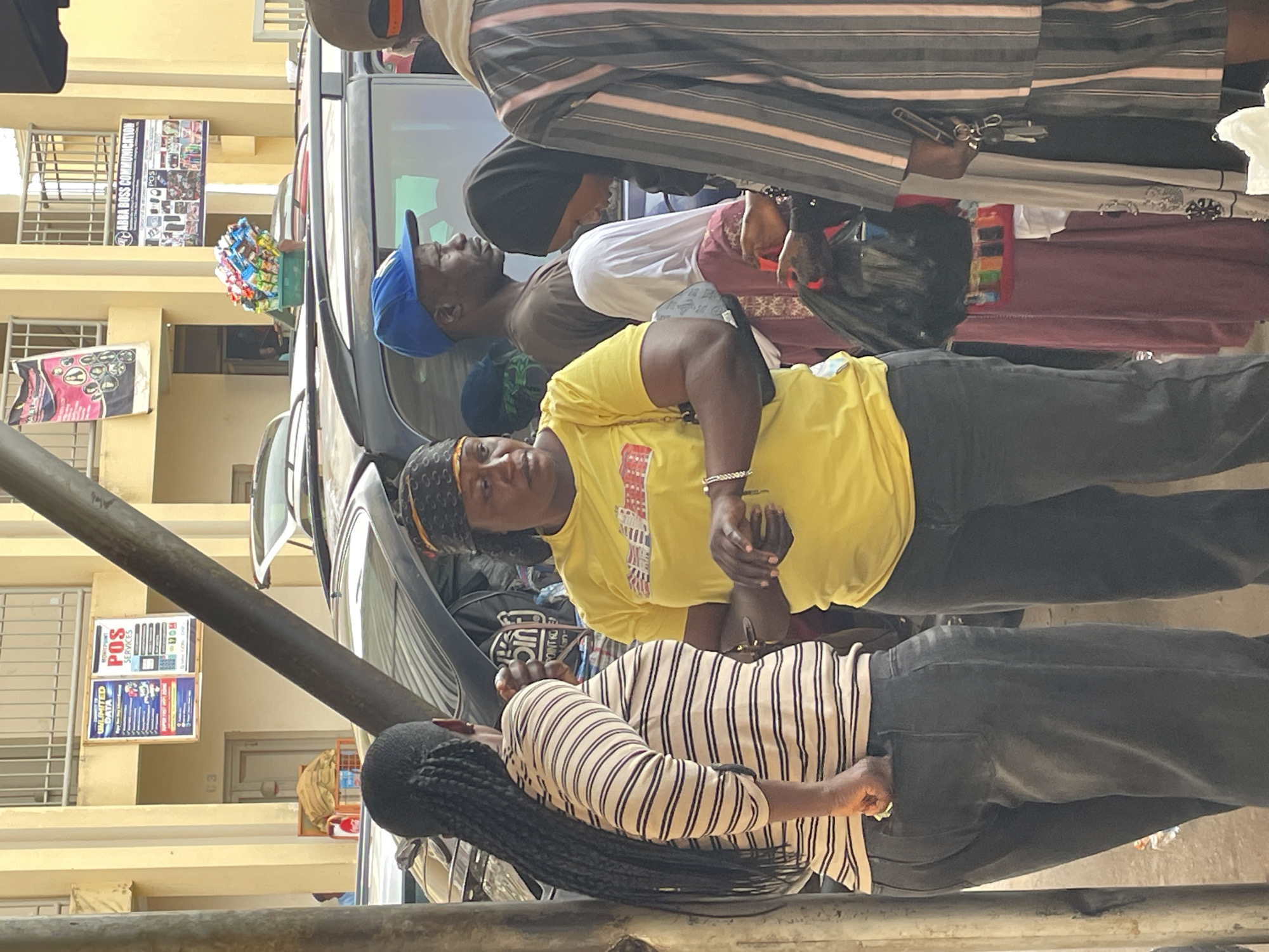 This image captures a vibrant street scene in Bukuru, Nigeria, featuring several individuals and commercial elements under bright daylight.
In the foreground, a woman in a bright yellow t-shirt and dark pants, wearing a patterned headband, is prominent, looking directly towards the viewer with a neutral expression. She has a silver bracelet on her left wrist. Partially obscured in front of her is another woman with long black braids, wearing a striped long-sleeve shirt and dark pants, with her hand in her pocket.
Behind these two women, a man wearing a blue baseball cap, a white t-shirt, and a brown top is visible, looking upwards. Further into the background, other individuals are present, including one person wearing a striped garment and another with a dark head covering, standing near a dark-colored car, possibly an SUV or van.
The car has a small crate filled with various packaged snacks and candies visible on its dashboard or window ledge, suggesting informal vending or a small market stall. A black metal pole or railing runs horizontally across the lower part of the frame, partially obscuring the foreground subjects.
The background consists of light-colored, possibly beige or yellow, buildings with barred windows. Several commercial signs are displayed on these buildings. One banner clearly reads "ABBA BOSS COMMUNICATION" and shows images of various electronics like phones and accessories. Another sign advertises "POS SERVICES" and "UNLOCKED DATA," with details about data plans (e.g., "1GB", "2GB") and potentially a telecom logo. A third, partially visible sign has the letter "H" on it.
The overall scene suggests a busy public area, likely a market or commercial street, during the daytime with clear weather.