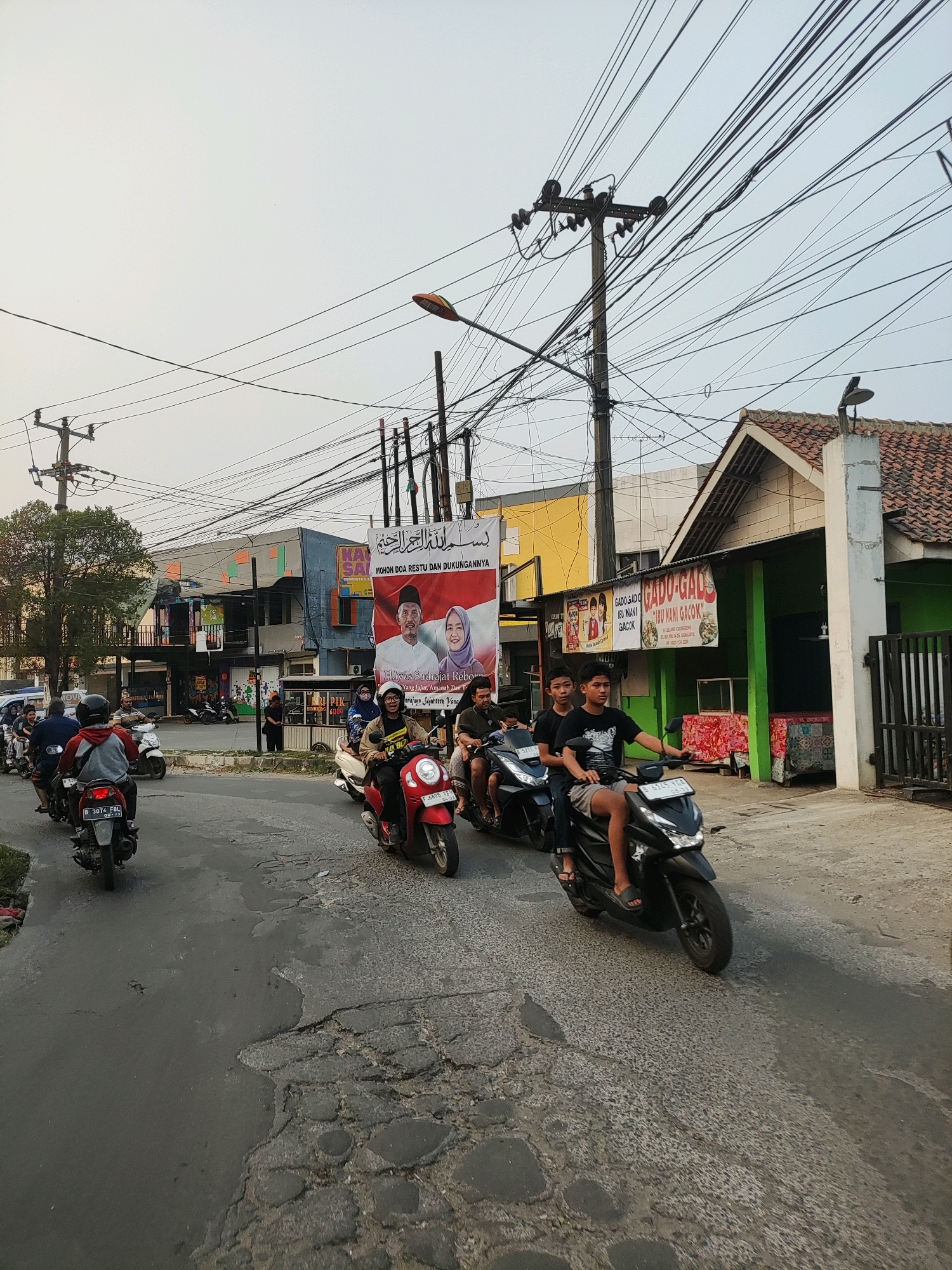 This is a full, eye-level shot of a street scene in Wanajaya, Indonesia, characterized by a procession of motorcycles on a cracked asphalt road. The sky is overcast and pale, suggesting early morning or late afternoon light. Overhead, a dense network of electrical wires crisscrosses the frame, supported by wooden utility poles. On the right side of the road, a green building with a red-tiled roof stands, featuring an awning with text that reads "GADO-GADO IBU NANI GACOK." A large banner with images of a man and a woman in formal attire, accompanied by Arabic script and Indonesian text, is prominently displayed on a building on the left.
Several people are riding motorcycles. In the foreground on the right, a young male is riding a black scooter, wearing a t-shirt and shorts, with two passengers behind him, also young males. To the left, a red scooter carries a rider wearing a yellow jacket and helmet, with a passenger. Further back, a procession of motorcycles continues, some with multiple riders. The road surface is heavily damaged, with numerous potholes and crumbling asphalt. Vegetation is visible on the far left of the image, with trees lining the road. The overall impression is of a busy, functional street in a local urban setting.