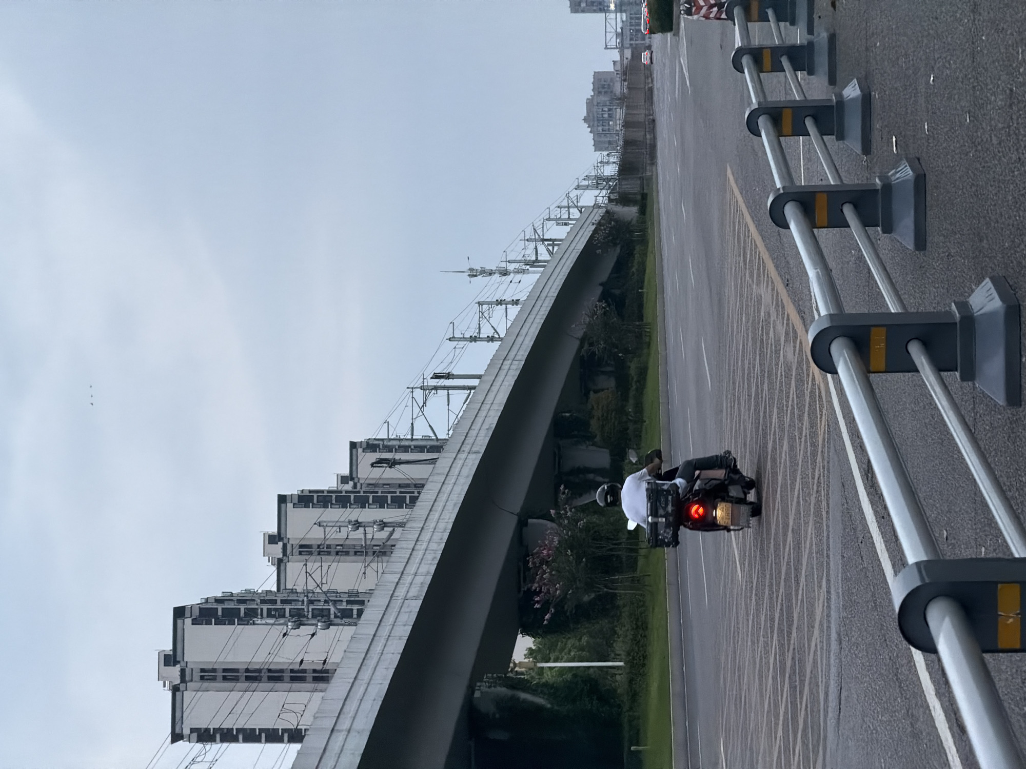 A person rides a scooter on a road in Jiangjin, China. The rider wears a white shirt and a black helmet, and the scooter has a black storage box on the back. The scooter is moving away from the camera. In the background, a large elevated structure, likely a railway or highway overpass, runs across the scene. Several tall apartment buildings are visible behind the overpass. The sky is overcast and a pale blue. The road has multiple lanes and is marked with yellow and white lines. A gray metal barrier with yellow markings separates the road from the sidewalk on the right side of the image. There are a few distant vehicles on the road.
