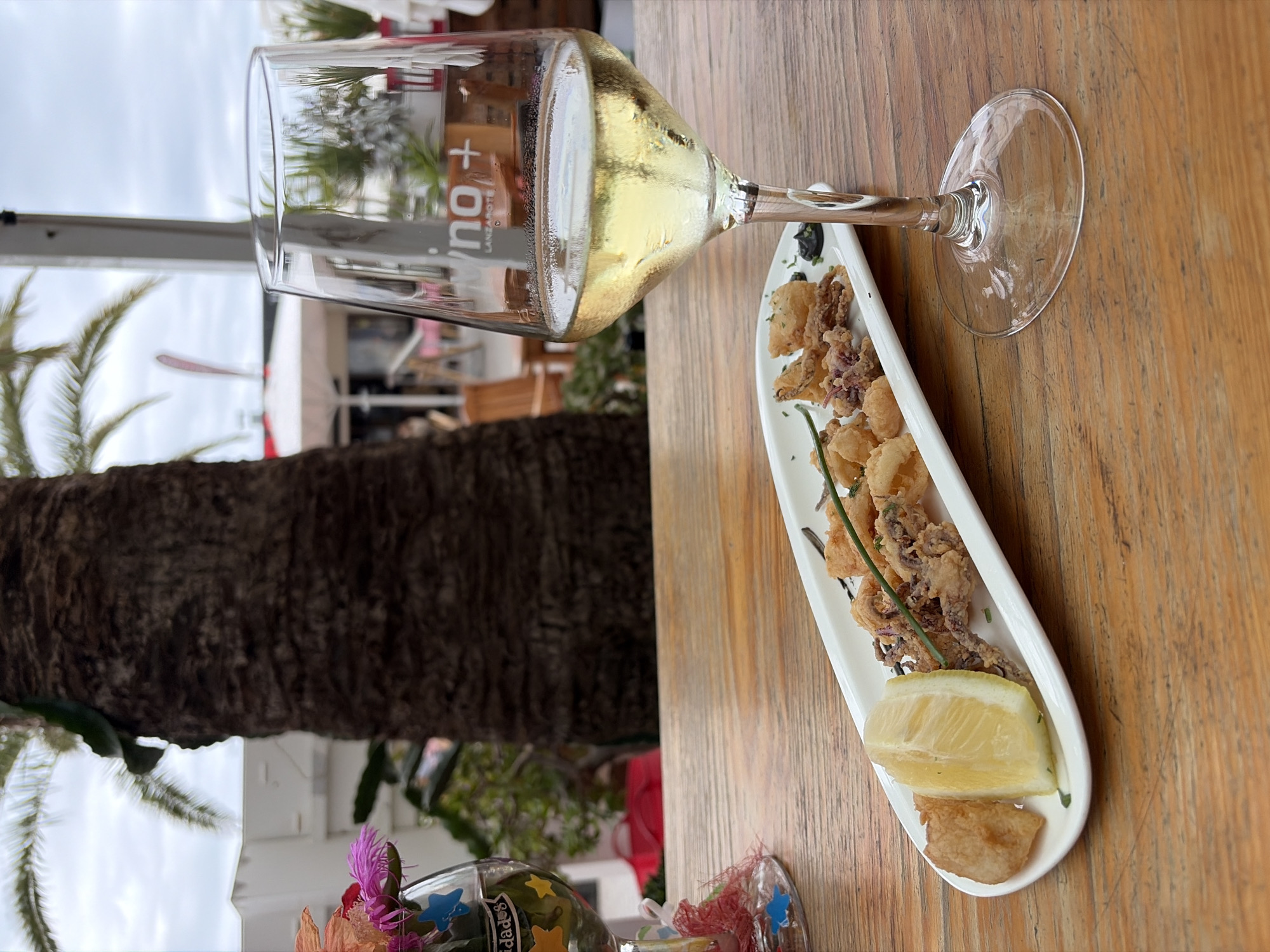 The image displays an inviting outdoor dining scene, likely in Puerto Del Carmen, Spain, given the "Lanzarote" inscription on a glass. In the foreground, a light-colored wooden table serves as the setting.
Prominently featured is a clear wine glass, partially filled with a chilled, pale yellow liquid, suggesting white wine or a sparkling beverage, indicated by the visible condensation on its surface. The text "wino+" and "LANZAROTE" can be seen on the side of the glass, viewed through the liquid.
Adjacent to the glass, an elongated white ceramic dish holds a serving of fried seafood, primarily crispy calamari and possibly other small fried pieces, garnished with green herbs. A fresh lemon wedge rests on the dish, ready to complement the seafood.
In the bottom left corner, part of a decorative item is visible—a small glass adorned with blue and yellow stars and pink, feathery embellishments. The Spanish word "felicidades" (congratulations) is faintly discernible on this item.
The background reveals an outdoor setting, dominated by a thick, dark brown palm tree trunk on the left. Beyond this, palm fronds, white structures (possibly buildings or tents), and an overcast sky suggest it is daytime. The overall ambiance evokes a sense of relaxation and leisure, indicative of a holiday or casual meal in a warm, coastal environment.