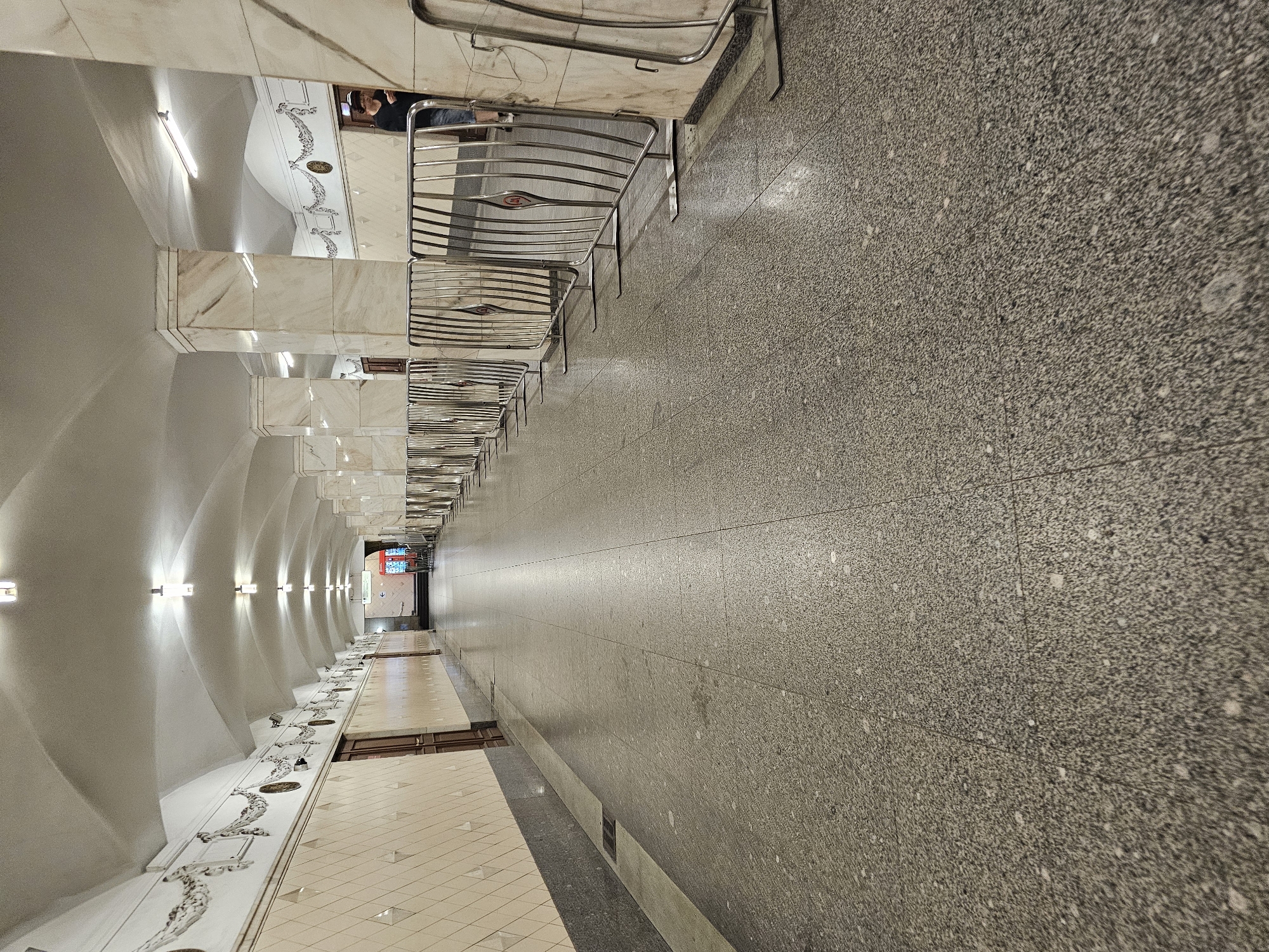 The image depicts an interior view of a long, arched hallway, likely within a Moscow Metro station. The hallway features marble-clad columns and walls decorated with patterned tiles and ornate friezes. A line of portable metal crowd control barriers runs along the right side of the hallway, separating the main walkway from the wall. The floor is made of polished terrazzo. At the far end of the hallway, an illuminated sign indicating direction is visible, along with red barriers. A single person wearing a dark top and blue shorts is partially visible standing near a doorway on the right side of the hallway, looking towards the far end. The scene is brightly lit by overhead fluorescent lighting.