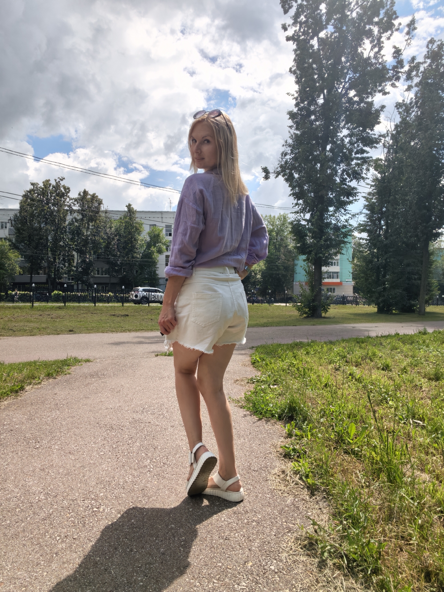 A young woman stands on a paved path in a park, looking over her shoulder at the camera. She is wearing a light purple shirt, white denim shorts with frayed edges, and white platform sandals. Her blonde hair is shoulder-length, and she has sunglasses perched on her head. The path curves to the left, bordered by green grass on the right and a darker paved area on the left. In the background, there are trees, buildings, and a cloudy sky. The sun casts a shadow of the woman on the path. The image is likely taken in the city of Sterlitamak, Russia, given the context.
