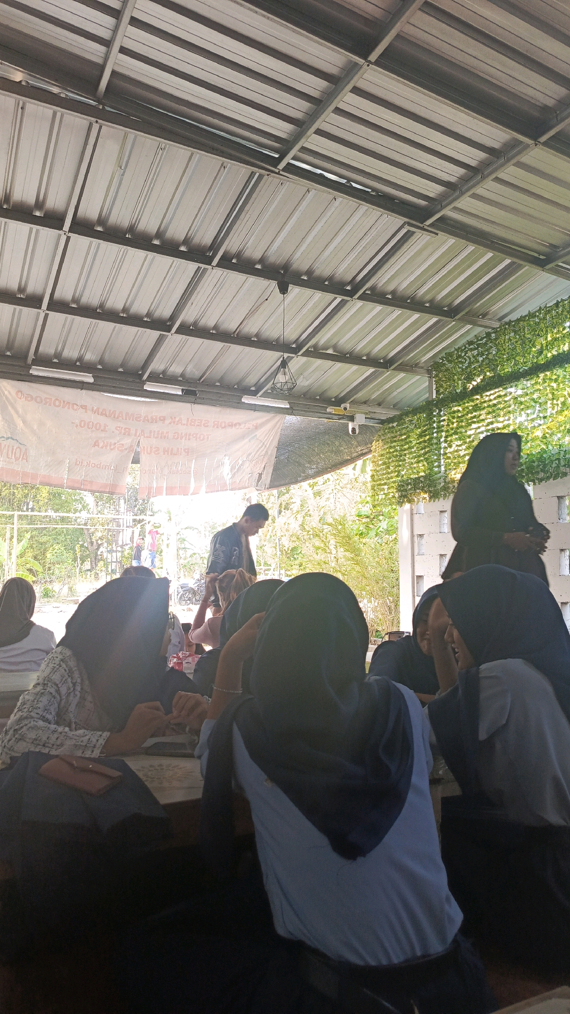 The image depicts an outdoor gathering, possibly a casual event or a religious meeting, taking place under a metal-roofed shelter. In the foreground, several individuals, predominantly women wearing hijabs, are seated around a table. Their faces are obscured by shadows or the angle of the shot, but they appear engaged in conversation or activities at the table, with some hands visible. A brown wallet is placed on the table in front of one of the seated individuals. In the middle ground, a young man in a dark jacket is walking away from the camera, with other people visible further in the background. A large banner with text in Indonesian hangs on the wall behind them, indicating a promotional or informational event. The environment suggests a warm climate, with lush green foliage visible in the background and sunlight filtering through the shelter. The scene is set in an open area, likely a community space or a park, given the presence of trees and a hint of a fence. There are no specific visual cues to pinpoint the exact location within Brahu, Indonesia, other than the general characteristics of an Indonesian outdoor setting.