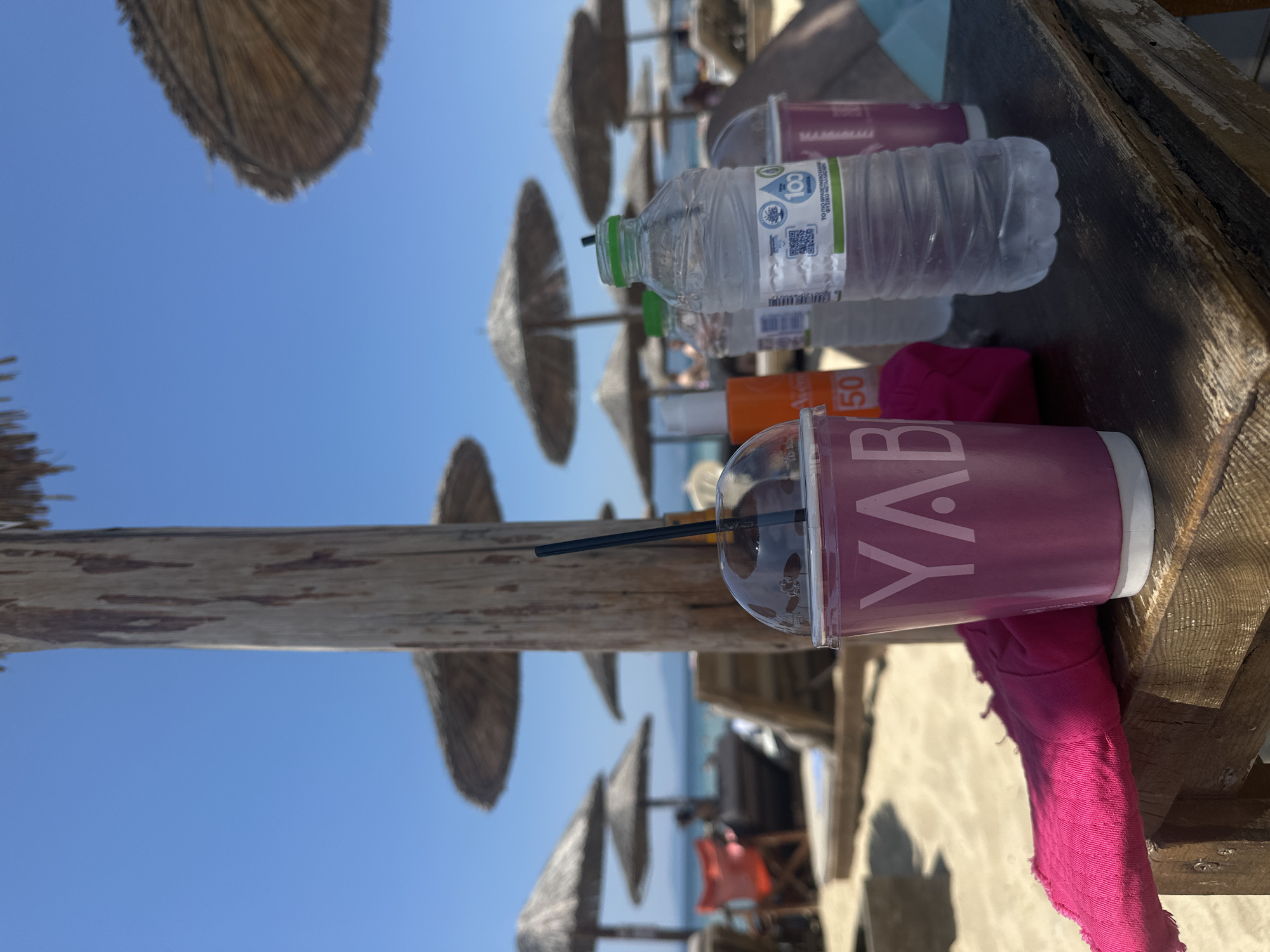 On a wooden table at Ofrynio Beach, Greece, several drinks are placed. In the foreground, a pink cup with a straw contains a dark liquid. Next to it, a bright pink cloth lies folded. To the right of the cup is a plastic bottle of water, its label featuring Greek text and a QR code. Behind the water bottle are two more cups, one purple and one pink, and an orange sunscreen bottle. The background shows a sandy beach with thatched umbrellas and lounge chairs, with the sea and a clear blue sky visible beyond.