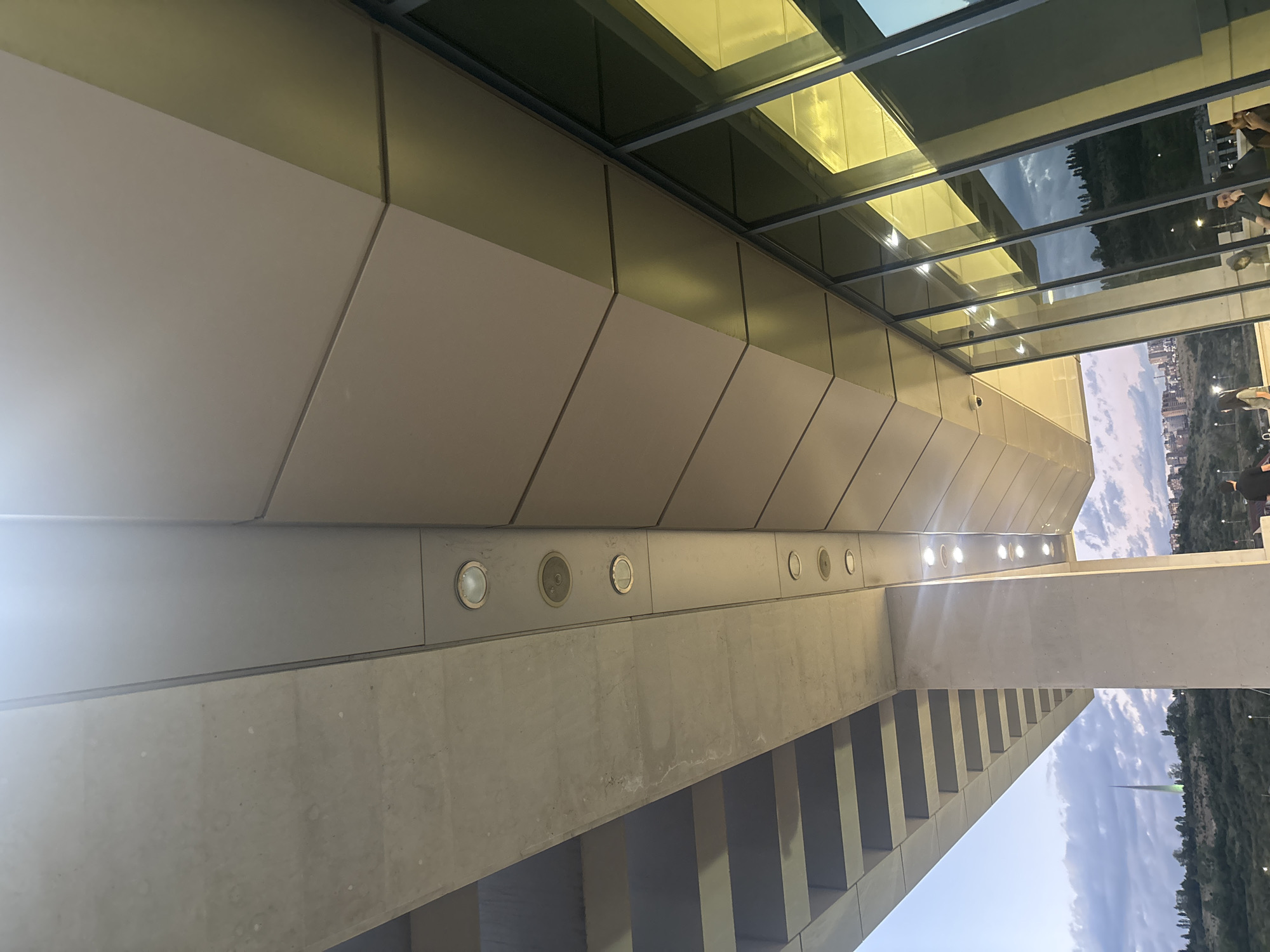The image displays a modern architectural structure, likely part of a building or a sophisticated walkway, captured from a low, upward-looking perspective. The overall scene suggests an urban setting against a natural backdrop.
The structure itself is characterized by striking geometric lines and a combination of materials. The primary façade visible on the left consists of light beige or off-white angular panels. Above these, a large section features a glass canopy or roof, reflecting the sky, distant green hills, and parts of a city. The reflections also show interior lights, casting a warm yellow or golden glow within the glass-enclosed space.
Horizontally across the mid-ground, a substantial concrete beam or wall runs across the frame. This concrete element houses several circular, recessed light fixtures, many of which are illuminated. Below this, a series of parallel concrete supports or steps descend downwards, forming a textured, ribbed pattern.
In the distant background, a hilly landscape covered with abundant green foliage is visible under a partly cloudy sky. Interspersed within the hills are glimpses of urban development, including buildings. A distinct green spire or antenna rises vertically from the distant horizon. A few indistinct human figures can be seen in the far right background, and their reflections are also present in the reflective glass panels.
The lighting suggests late afternoon or early evening, with a bright source creating a lens flare on the left. The scene is located in Yerevan, Armenia, as indicated by context. No visible text is present.