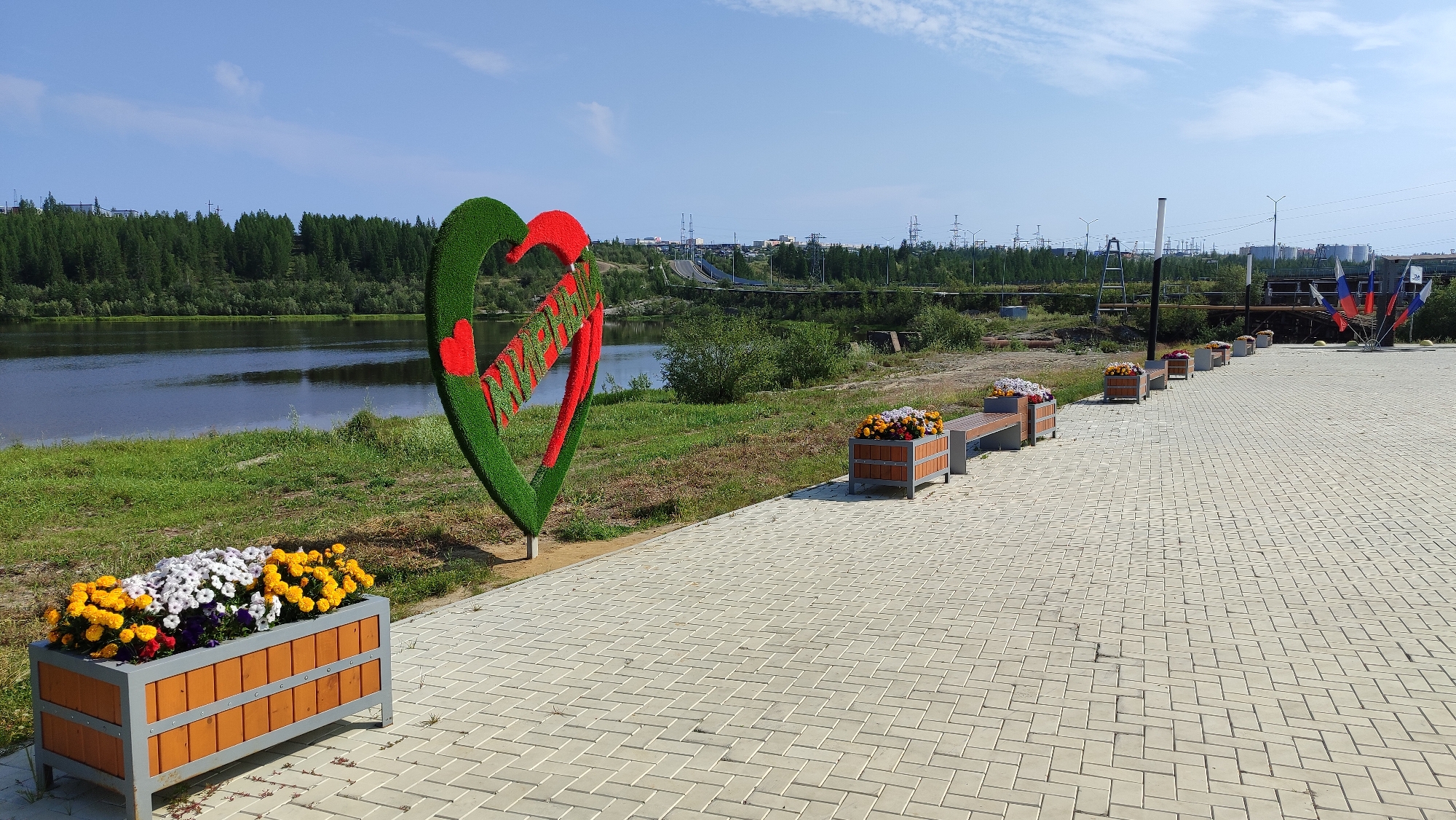 This image depicts a paved walkway alongside a river in the city of Mirny, Russia. On the left side of the walkway, a large, green, heart-shaped sculpture adorned with red accents is prominently displayed. In front of this sculpture, a wooden planter box is filled with colorful flowers, including yellow, white, and purple blooms. Further down the walkway, several more wooden planter boxes and benches are arranged along the path. Beyond the walkway, a wide river flows, with a lush green bank and a dense forest covering the opposite side. In the background, a distant cityscape is visible, characterized by industrial structures, power lines, and buildings. The sky above is clear and blue with a few scattered clouds. A cluster of Russian flags is planted on the right side of the walkway in the distance. The overall scene suggests a well-maintained public area, possibly a park or promenade, with elements of nature and urban development coexisting.