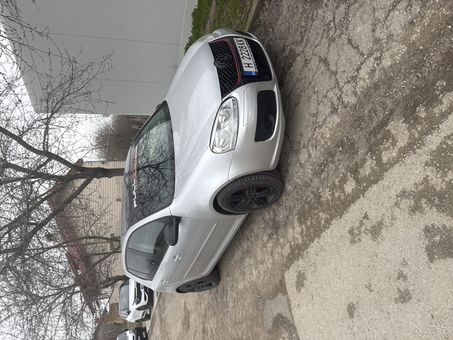 A silver Volkswagen Golf Mk5 hatchback is parked on an unpaved dirt and gravel surface, adjacent to a patchy asphalt road, in the city of Stan, Bulgaria. The vehicle features black multi-spoke aftermarket wheels, revealing red brake calipers, and has a black roof. A black sun visor strip on the windshield displays white text, partially legible as "FULL PERFORMANCE." The front grille incorporates a red accent line, and the Bulgarian license plate "H 2228 KK" is mounted below. In the background, to the left, a two-story house with light-colored siding and a red-tiled roof is visible, with other parked vehicles, including a white van and a white SUV, further down the road. To the right, a large, plain, light-colored building stands. Several deciduous trees with bare branches are scattered across the scene under an overcast sky, indicating a winter or early spring setting. No individuals are present.