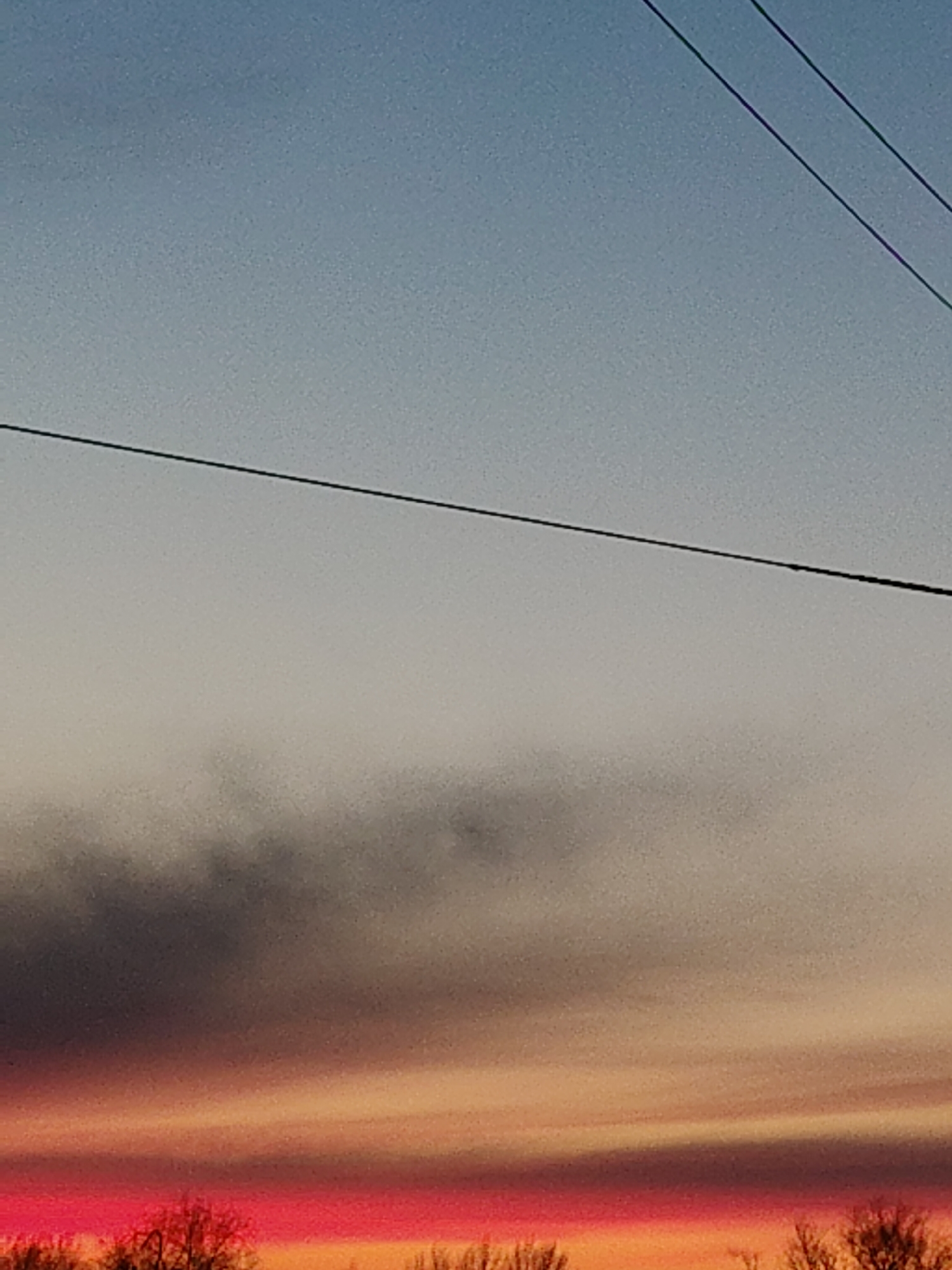 The image depicts a sky at dusk or dawn, showing a gradient of color from blue at the top to shades of gray, orange, and pink near the bottom horizon. A dark band of clouds is visible in the middle. Silhouetted tree branches appear along the bottom edge. Power lines stretch across the upper portion of the frame. The location is identified as Unknown, Unknown.