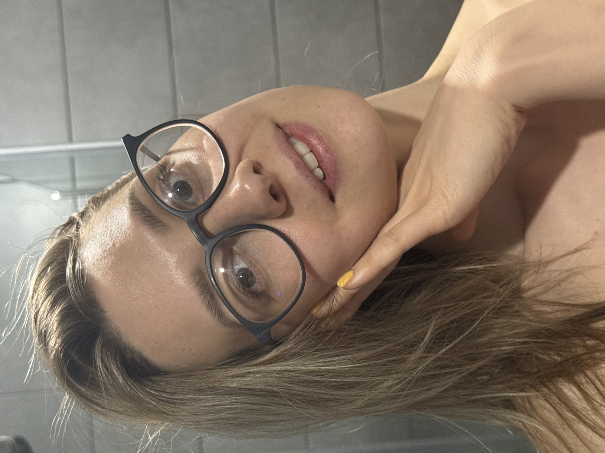 A woman wearing glasses is photographed from a low angle, her face tilted to the side. She has fair skin, light brown hair, and is wearing a yellow nail polish. Her right hand is positioned near her face. The background appears to be a plain gray wall or surface. The location is not discernible from the image.