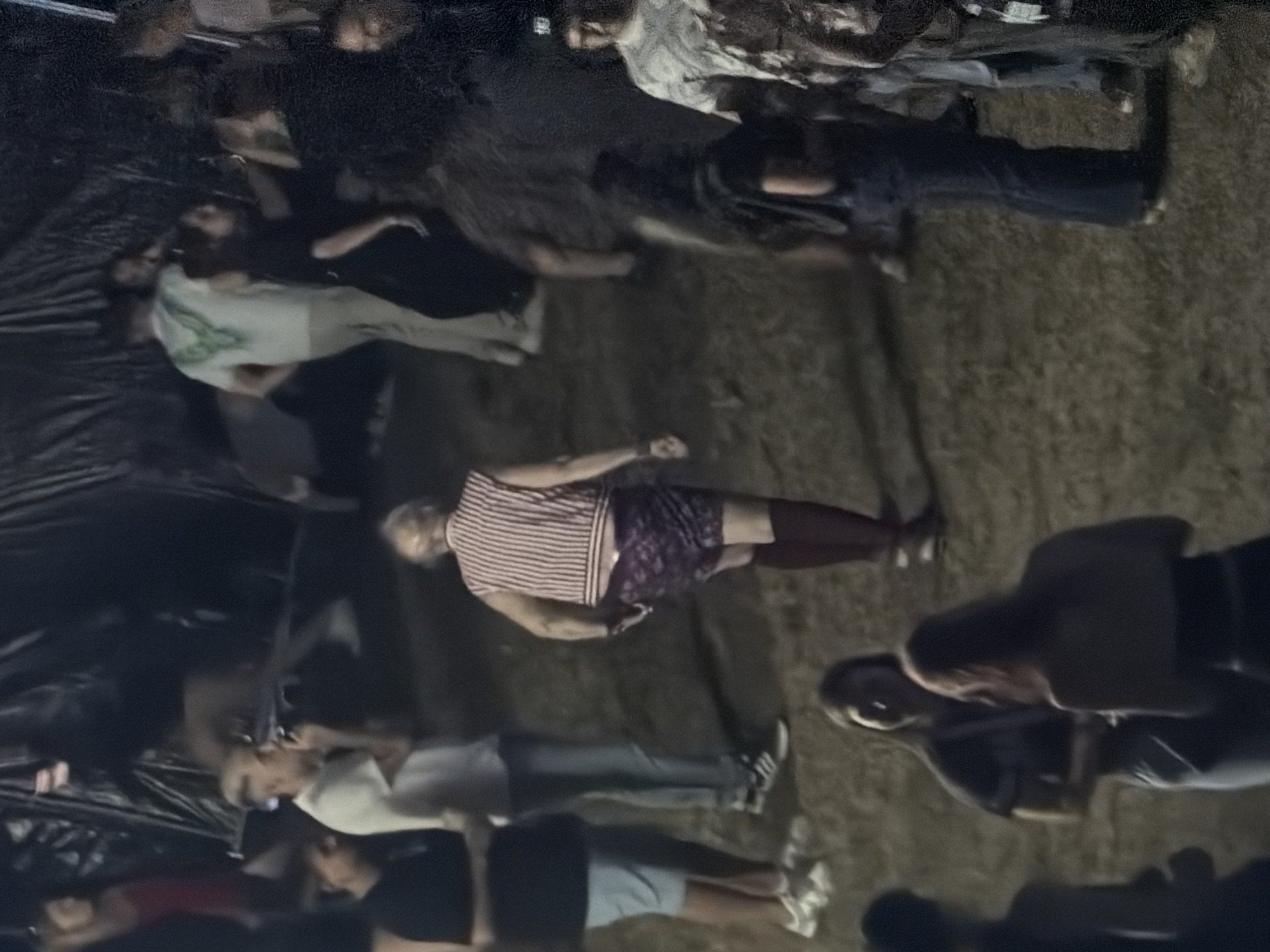 The image depicts a gathering of people outdoors at night. In the foreground, several individuals are visible, some facing the camera and others with their backs turned. The central figure is an older person wearing a striped vest, patterned shorts, and dark socks, walking away from the camera. To the left, a person in a black t-shirt and light blue shorts stands beside another in a white t-shirt and jeans. Further back, a group of people are indistinctly visible, including one wearing a white t-shirt with a green design and another in a dark dress. The ground appears to be covered in dirt or grass, and the background is dark with some structures partially visible, suggesting an outdoor event or venue. The overall lighting is dim, creating a low-visibility environment typical of nighttime photography. There is no specific indication of the city of Echternach, Luxembourg in the image content.