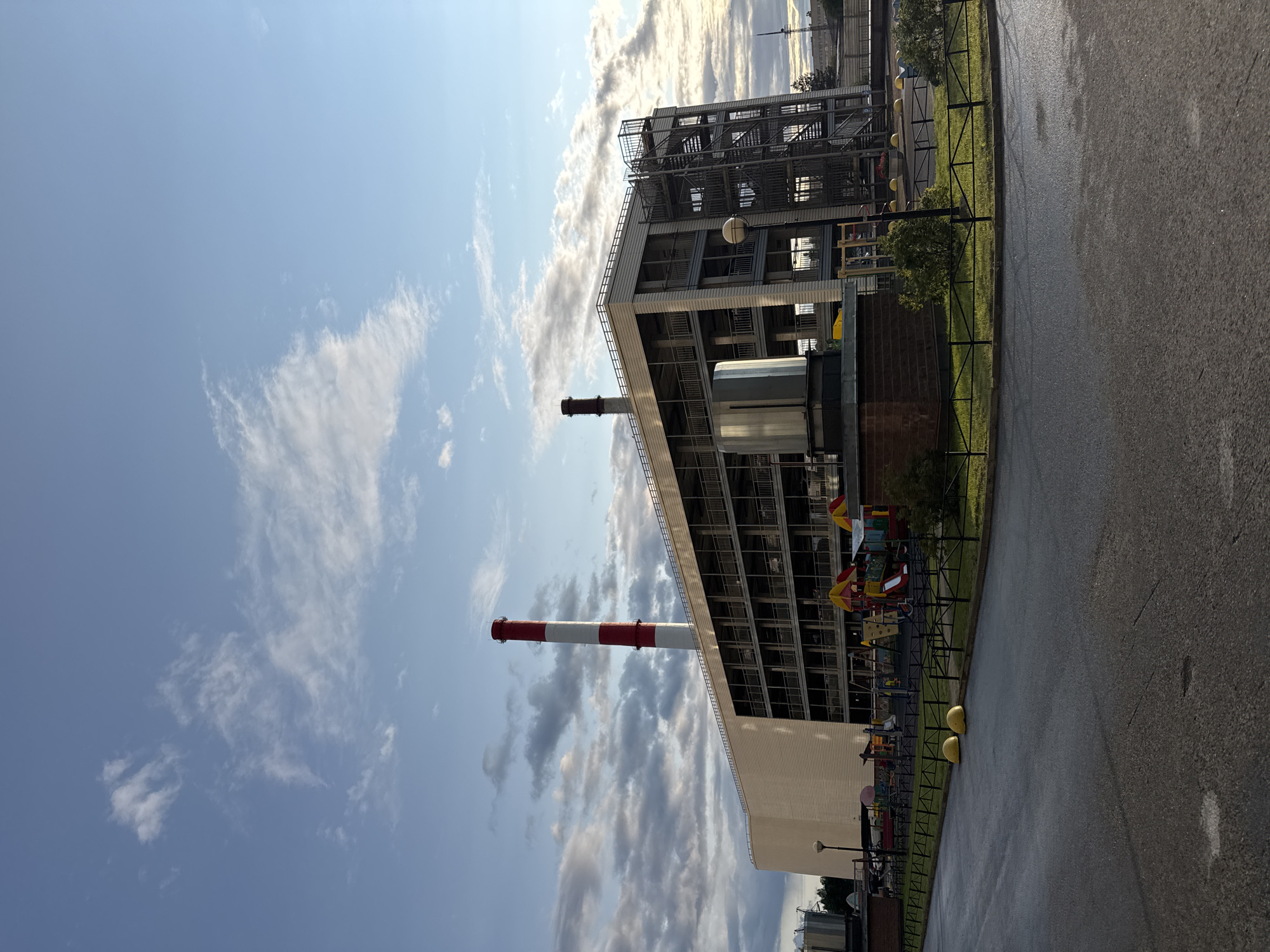The image shows a multi-story building with an industrial appearance, possibly a parking garage or a facility with multiple levels. It features prominent red and white striped smokestacks on the left side of the building. There is a metal staircase on the right side of the building, indicating access to different levels. In front of the building, there is a children's playground with colorful equipment. The area appears to be an outdoor setting, with a paved road or lot in the foreground and a patch of grass with a fence. The sky is partly cloudy. Based on the provided information, this scene is located in the city of Sinyavino, Russia. There are no people visible in the image.