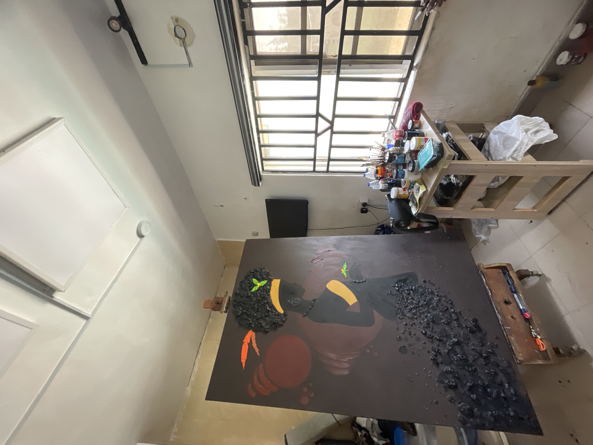 The image shows an art studio in a residential building in Ado, Nigeria. The room has white walls, a window with metal bars, and a concrete floor. An easel supports a large canvas depicting a stylized portrait of a Black woman with a brown background and orange and green accents. Art supplies, including paints and brushes, are organized on a wooden shelf near the window. Additional materials, such as a black speaker and bags, are scattered around the floor near the canvas and shelf. There are no people visible in the image.