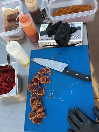 The image depicts a culinary preparation scene. A blue cutting board dominates the lower center of the frame, upon which cubed pieces of seared tuna, coated with sesame seeds, are arranged. A chef's knife with a black handle rests near the tuna. A pair of black gloves are present, one worn on a hand visible at the bottom right corner of the frame, the other resting on a box of Konnoss XL nitrile gloves. Several containers are arranged around the cutting board: one holds cubed red produce (possibly tomatoes), another contains cubed brown food items, and three squeeze bottles containing sauces of different colors are positioned to the upper left. A container holding brownish granules is located in the upper right. The scene is likely set on a stainless steel table, potentially in a food preparation area. There is no explicit evidence to confirm the exact location as Barbate, Spain.