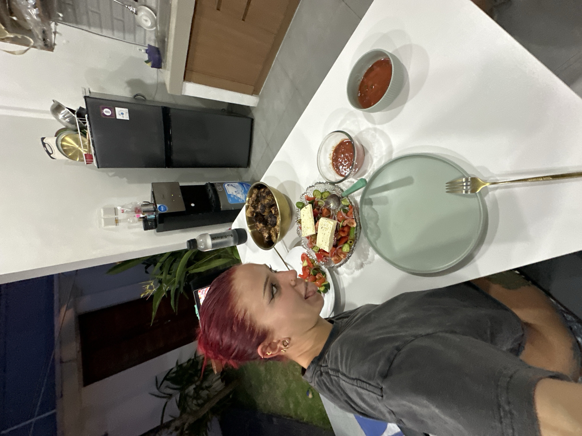 A woman with short, dyed red hair is sitting at a white table laden with food. The table contains a green plate with a gold fork, a salad with feta cheese, a bowl of stewed meat, and two smaller bowls of red sauce. The woman is wearing a grey t-shirt and appears to be about to eat. In the background, there is a kitchen area with a small refrigerator, cooking utensils hanging on the wall, and cabinets. Outside the kitchen, there is a glimpse of an outdoor area with greenery. The location is in North Kuta, Indonesia.