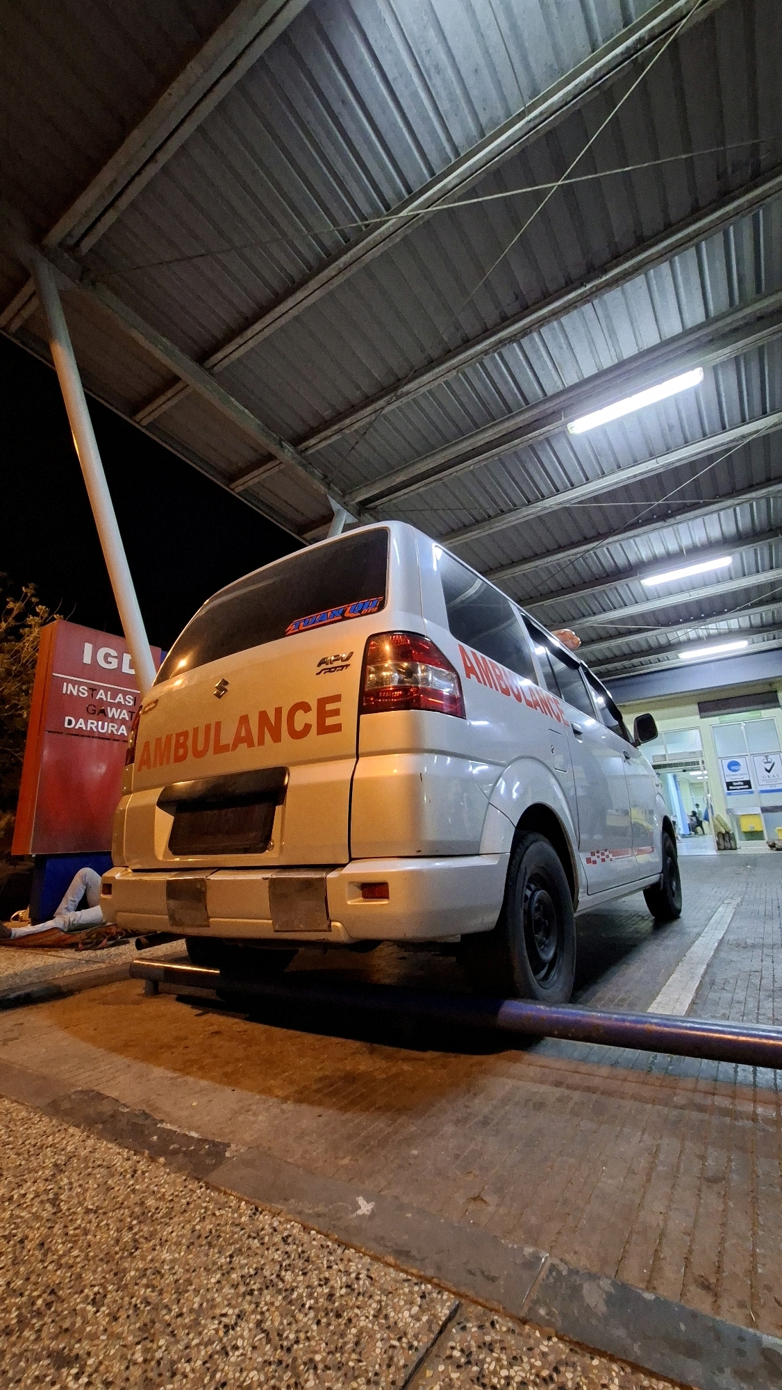An ambulance is parked under a metal roof structure at what appears to be a medical facility. The ambulance is white with red lettering that reads "AMBULANCE" on the rear and side. A red sign with white text that says "IGL INSTALASI GAWAT DARURA" (Emergency Care Installation) is visible to the left of the ambulance. A person in light blue pants and a white shirt is lying on the ground to the left of the sign, partially obscured by the ambulance. The ground is paved with concrete and speckled with gravel. Fluorescent lights are visible on the ceiling of the structure, and the scene is illuminated by artificial lighting, indicating it is nighttime. In the background, the entrance to a building with glass doors and medical signage is visible. The location is identified as Banda Aceh, Indonesia.